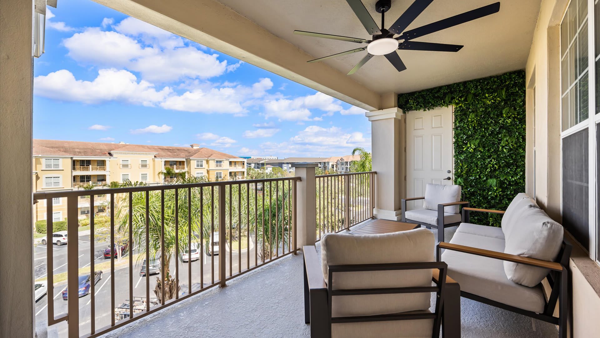 Expansive covered balcony with a ceiling fan and a green wall, featuring outdoor seating and offering a wide view of the residential complex and sky.