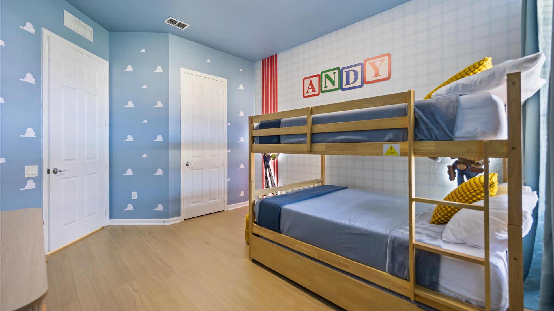 A wide view of the Toy Story themed bedroom, showcasing a light wood bunk bed with blue bedding and two white doors. The light blue walls are adorned with white cloud decals and a large 'ANDY' sign above the bed.