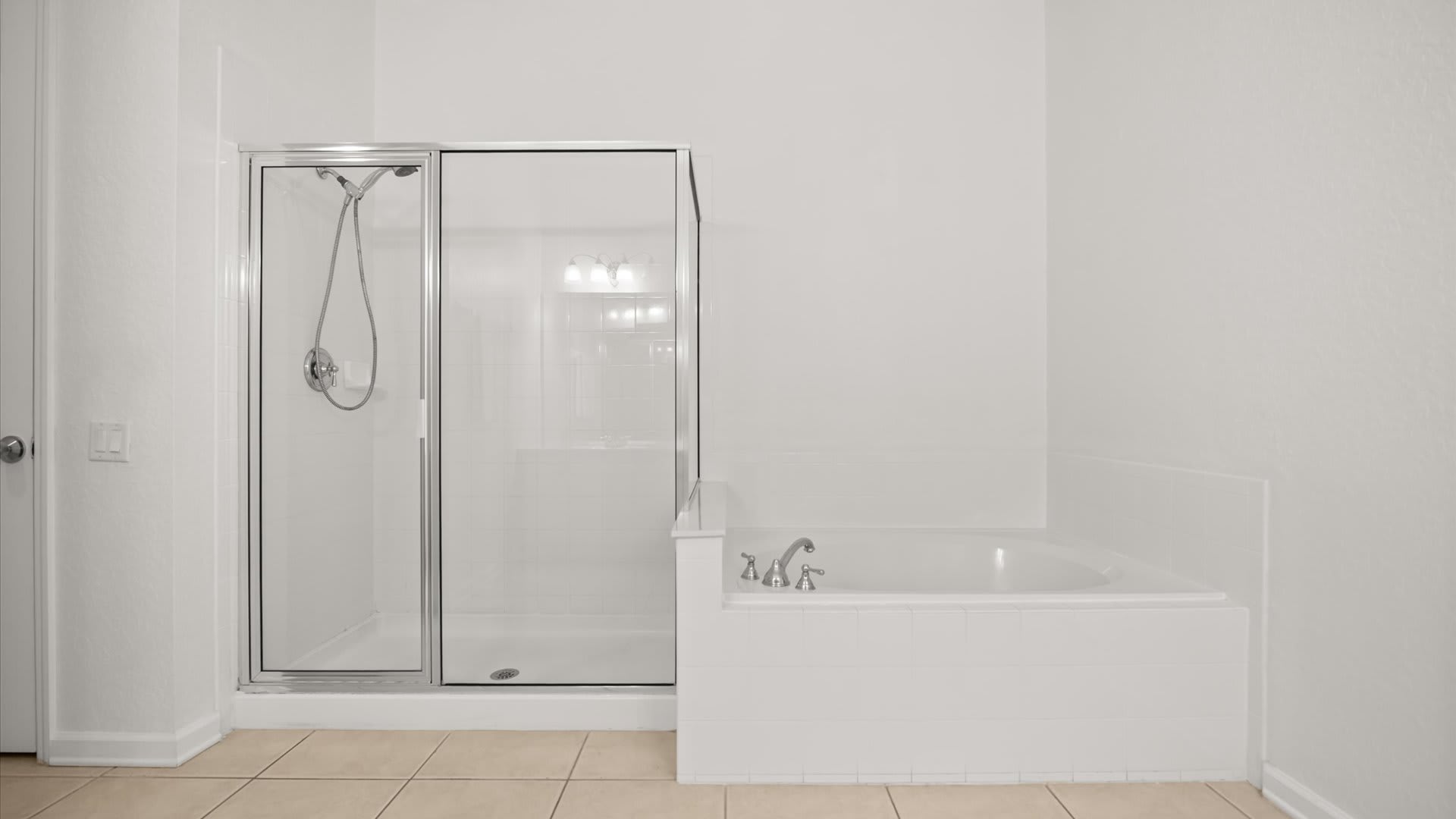 A bright white bathroom featuring a glass-enclosed shower stall with a handheld showerhead and a built-in corner bathtub with a white tiled surround. The floor is light-colored tile.