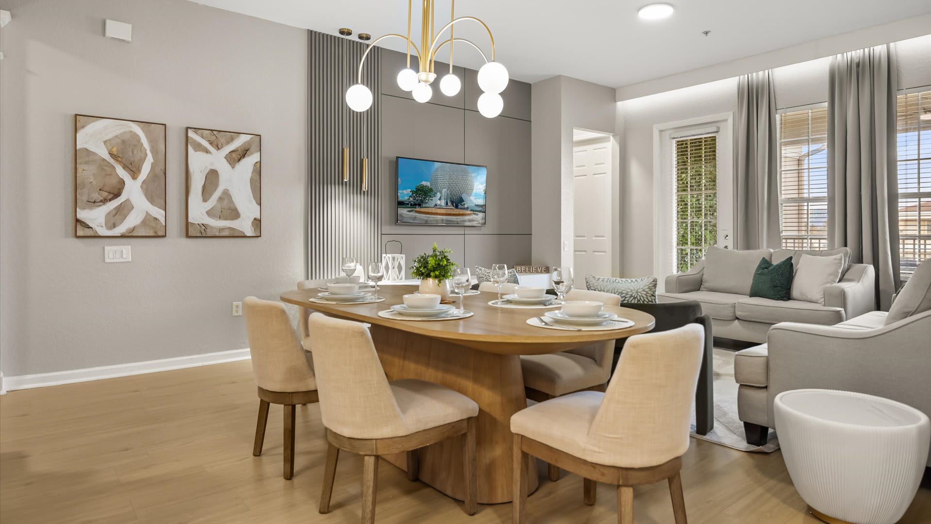 Modern dining area with a light wood oval table set for six, featuring cream upholstered chairs and a contemporary gold chandelier. A wall-mounted TV is visible on a gray accent wall, with abstract art on the adjacent wall.