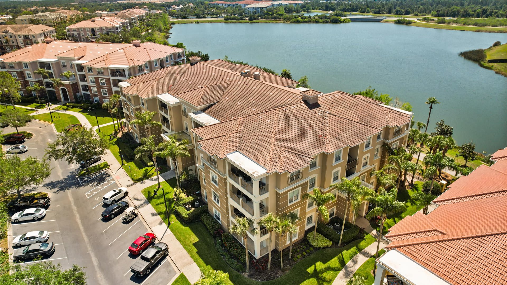 Aerial view of a large, multi-story condominium building with a terracotta tile roof and light-colored facade, showing multiple balconies and a parking lot with cars. A large lake is visible in the background, surrounded by green landscape.