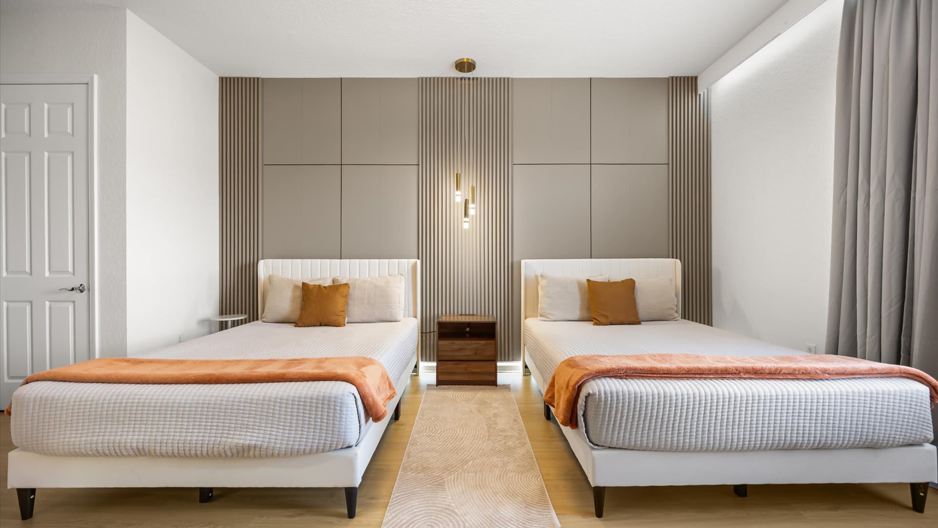 Modern bedroom featuring two single beds with white frames and orange throws, separated by a wooden nightstand. A textured gray accent wall with vertical slats and a gold pendant light fixture adds a stylish touch.