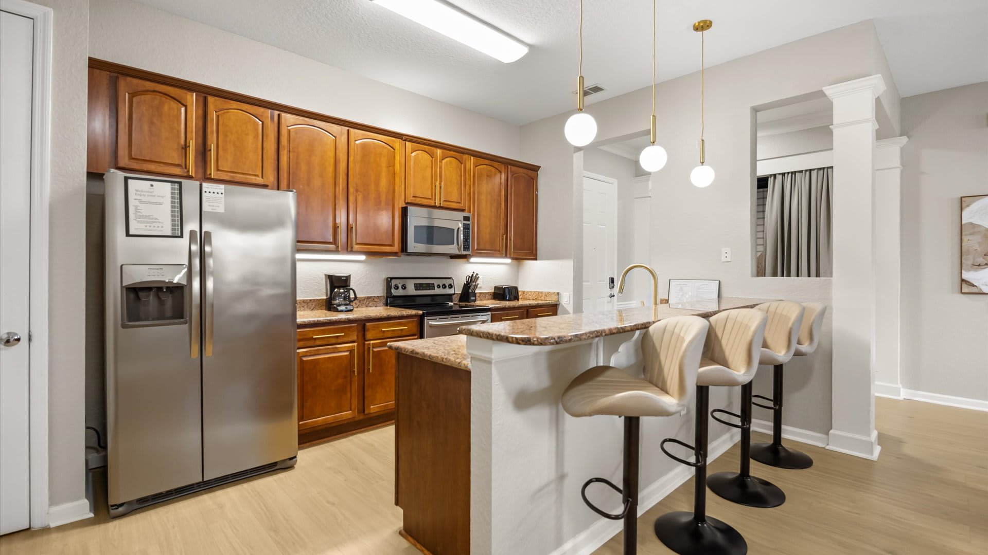 Kitchen with traditional wooden cabinetry, granite countertops, and stainless steel appliances. A breakfast bar with three light-colored bar stools and modern pendant lights separates the kitchen from the living area.