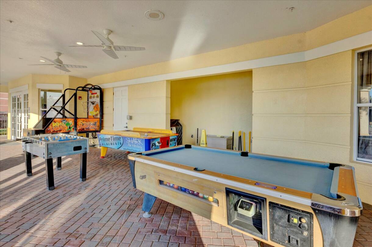 Outdoor covered game area featuring a pool table, foosball table, and arcade games on a brick-paved floor. The space is open-air with ceiling fans and light yellow walls.