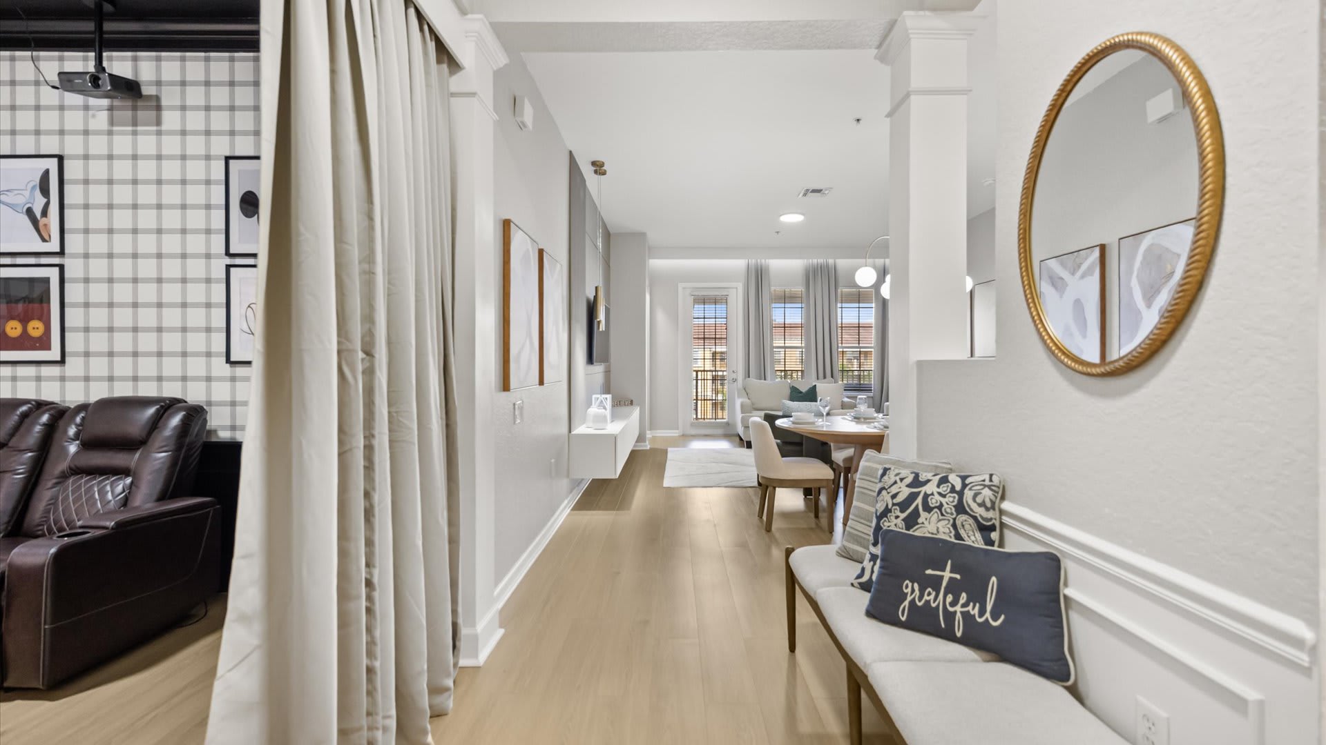 Hallway leading from a themed media room to the open-concept living and dining area, featuring light hardwood floors and a decorative bench.