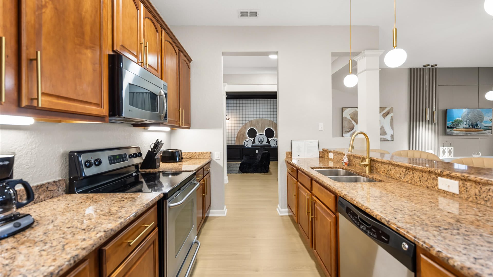 Galley-style kitchen with traditional wooden cabinetry, granite countertops, and stainless steel appliances. A view through to another room with Mickey Mouse decor is visible at the end of the kitchen.