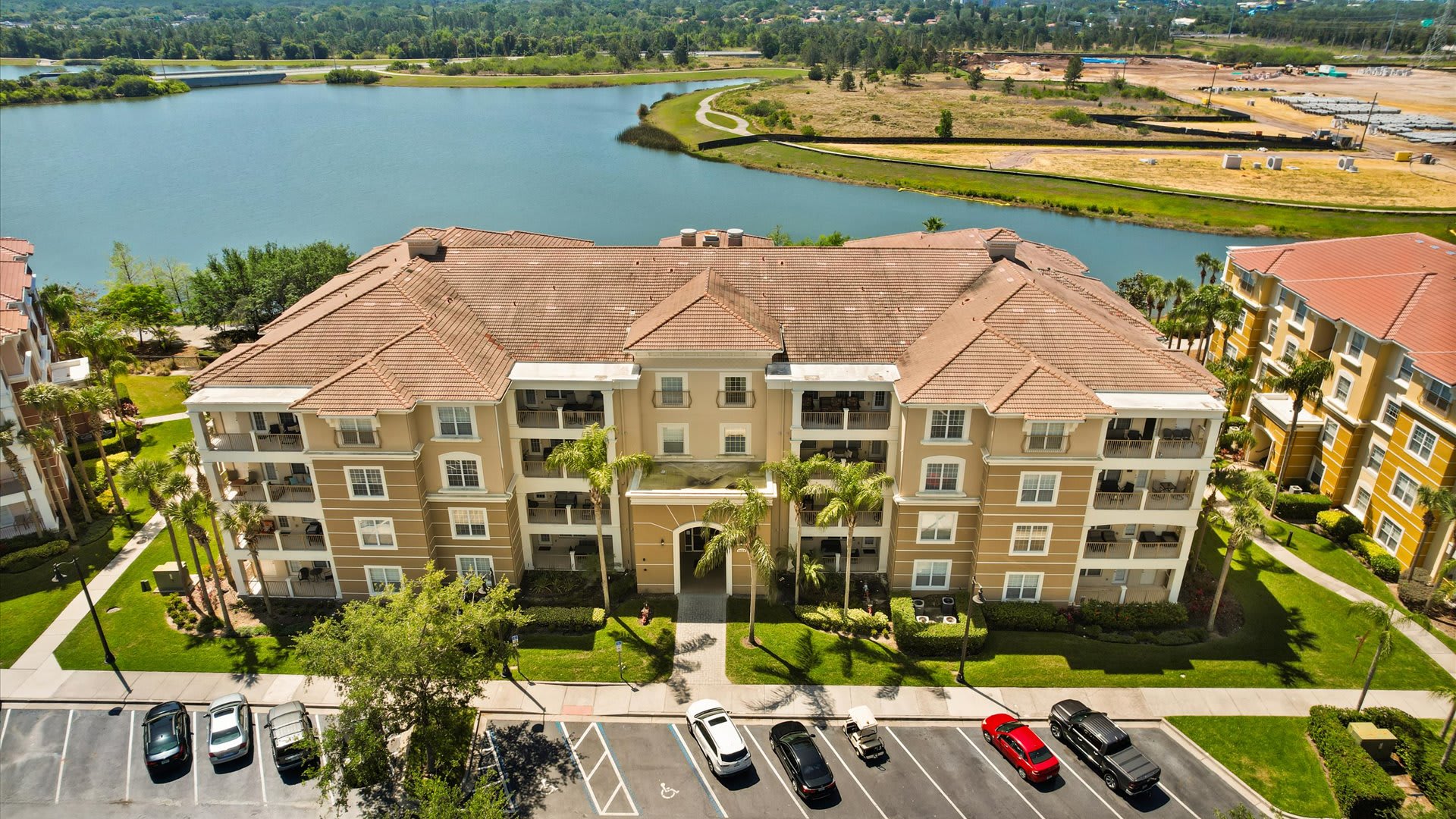 Aerial view of a large, multi-story condominium building with a terracotta tile roof and light-colored facade, featuring an arched entrance and balconies, with a parking lot in the foreground and a lake in the background. Palm trees line the building's perimeter.