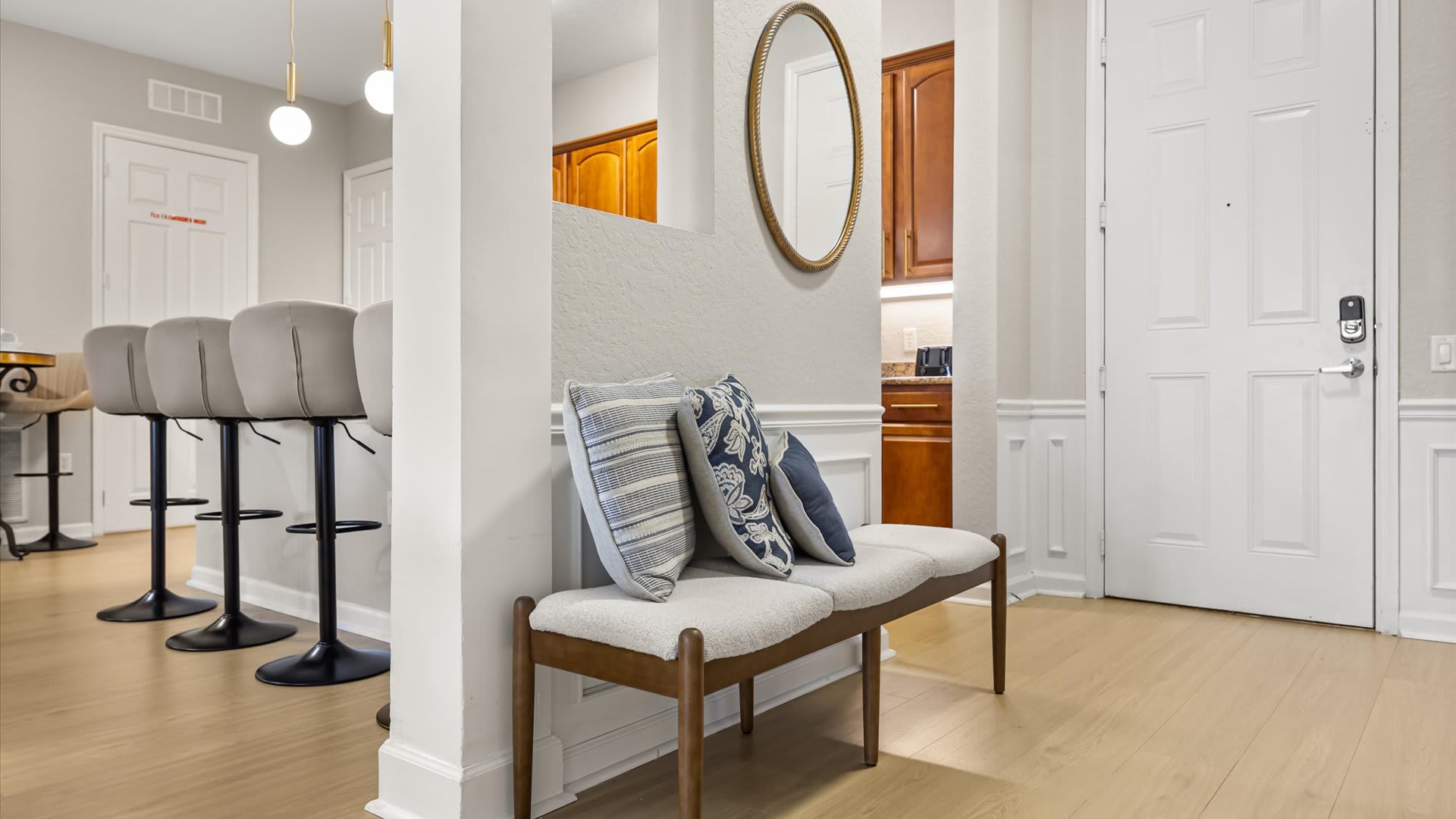 Inviting entryway with an upholstered bench adorned with throw pillows, a large oval mirror, and a view towards a bar area with high stools.