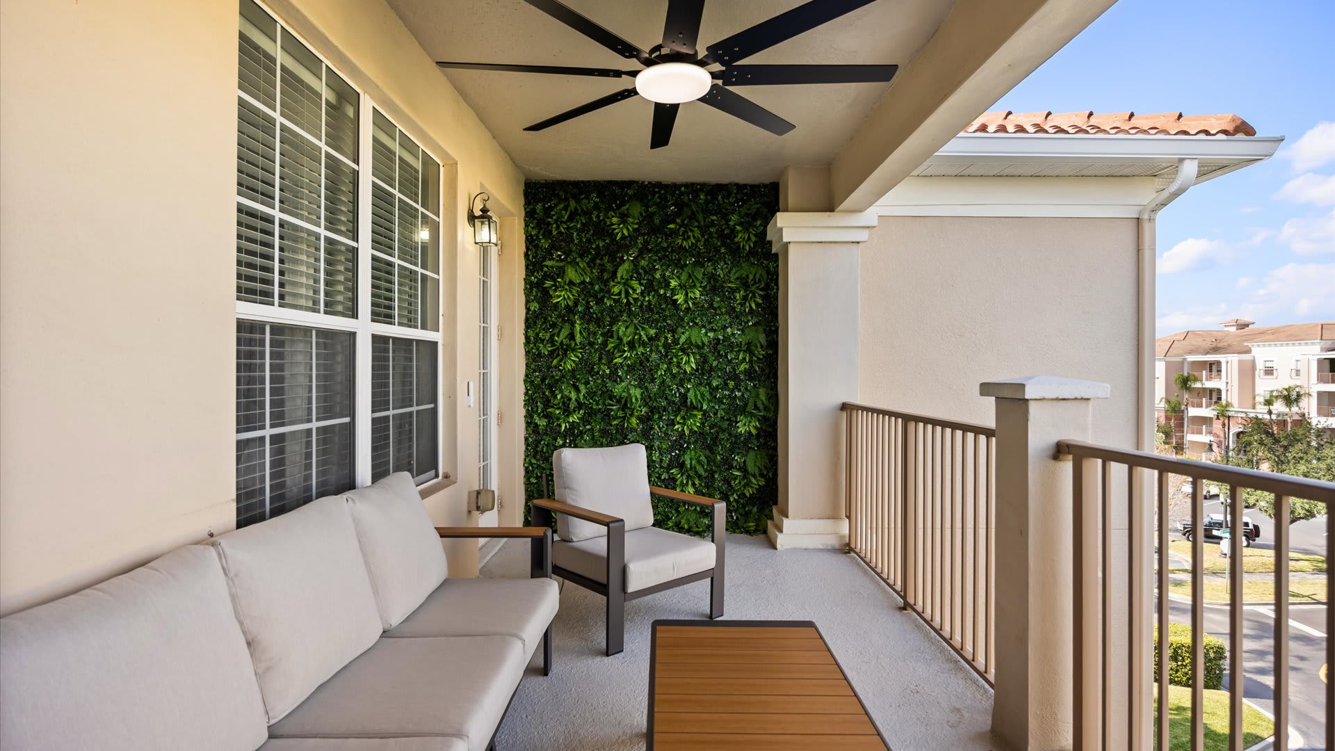 Covered balcony with a green wall feature, furnished with a light gray outdoor sofa, an armchair, and a coffee table, offering views of the surrounding buildings.