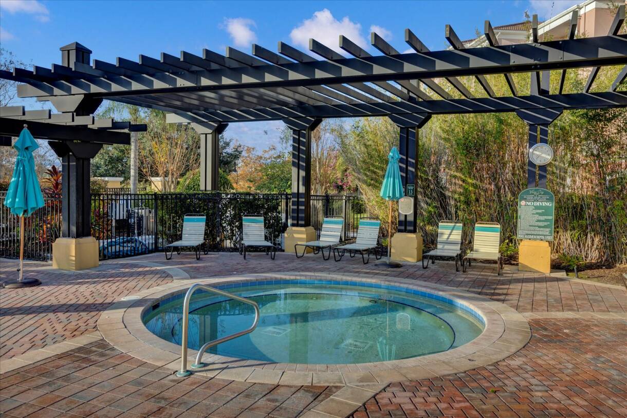 Outdoor hot tub area with a circular hot tub surrounded by brick pavers, lounge chairs, and a dark pergola providing shade. Teal umbrellas add a pop of color, and a "NO DIVING" sign is visible.