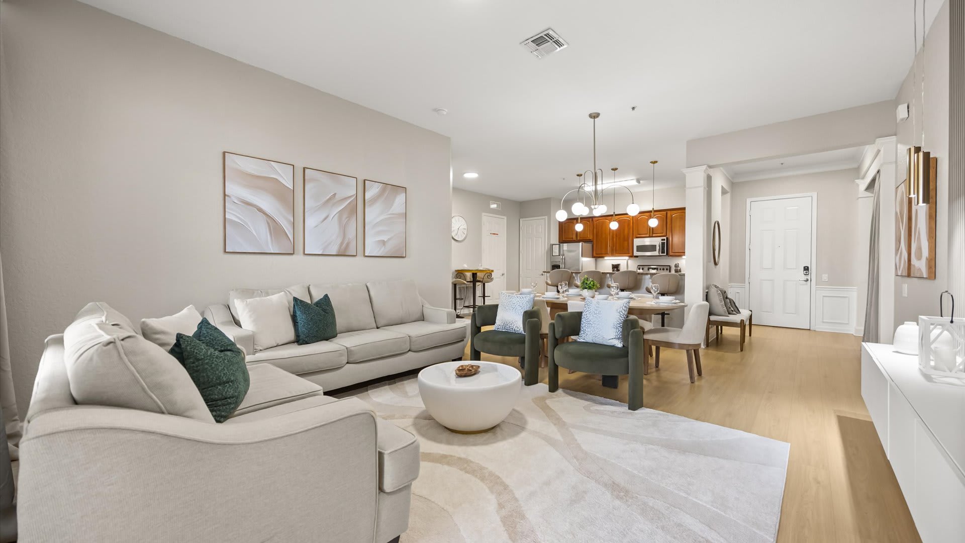 Wide view of the open-concept living room featuring a large L-shaped light gray sofa, a round white coffee table, and two dark green armchairs. Abstract art adorns the wall, and the dining area and kitchen are visible in the background.