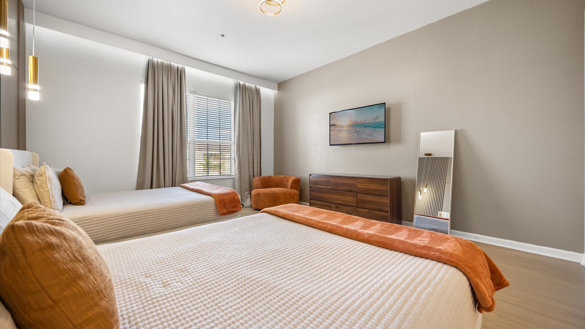 Modern bedroom with two single beds, each with white bedding and an orange throw. A wooden dresser with a wall-mounted TV above it, a full-length mirror, and an orange armchair are also visible.