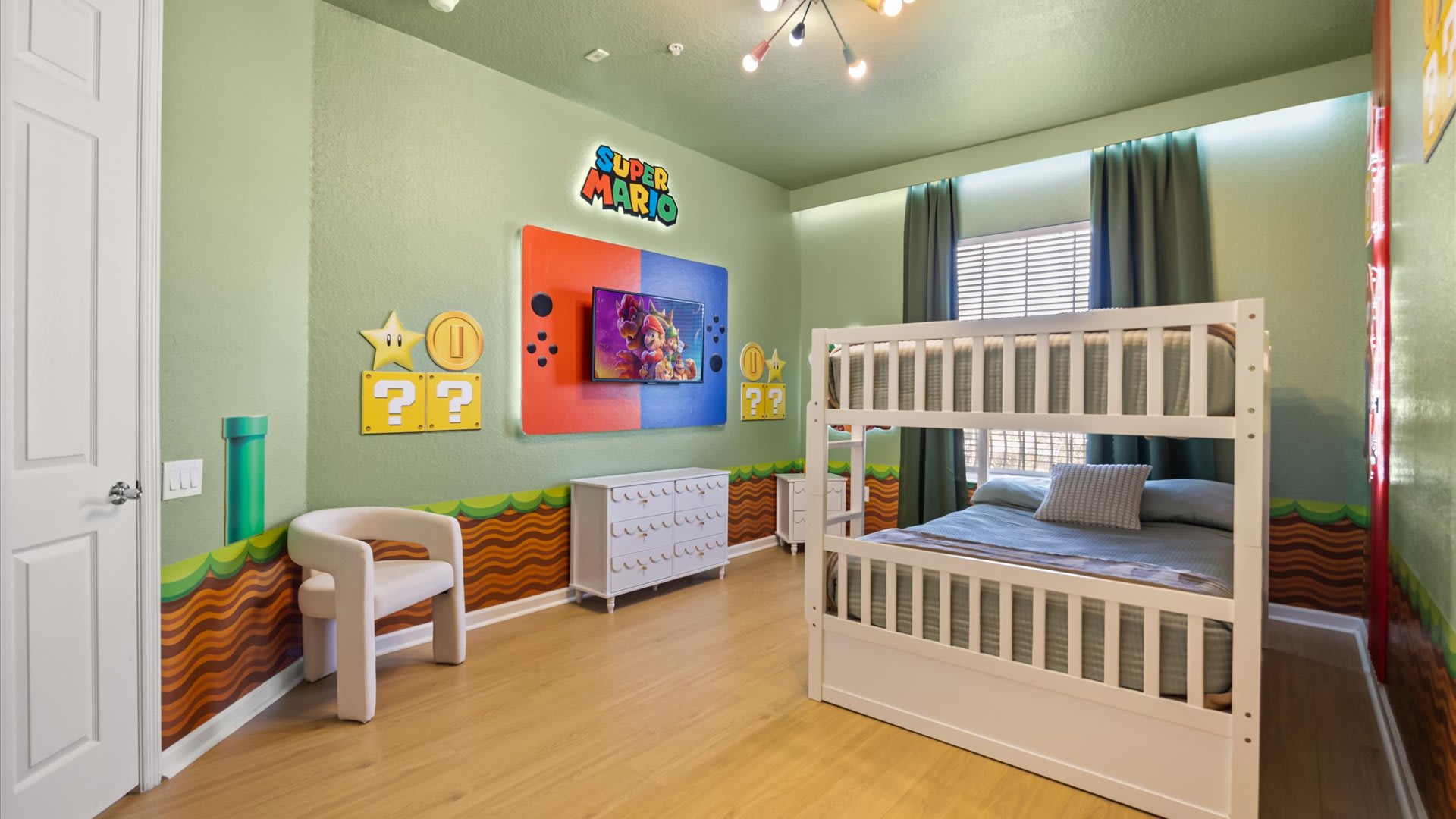 A wide view of the Super Mario themed bedroom, featuring a white bunk bed and a wall-mounted TV displaying a game. The light green walls are adorned with Mario-themed decals and a 'Super Mario' sign above the TV.