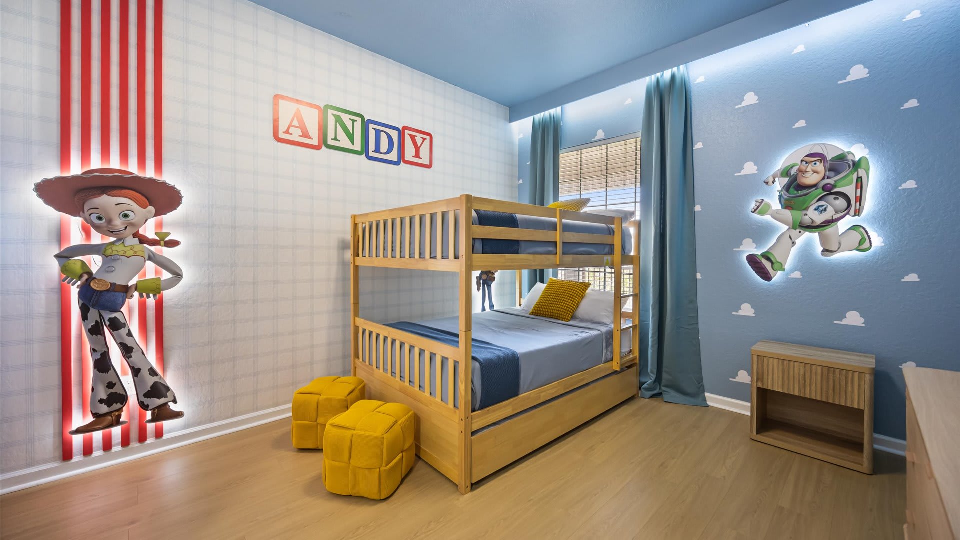 A vibrant Toy Story themed bedroom with a light wood bunk bed, blue bedding, and yellow poufs. The walls are light blue with white cloud decals, featuring large illuminated cutouts of Jessie and Buzz Lightyear, and 'ANDY' spelled out in colorful blocks above the bed.