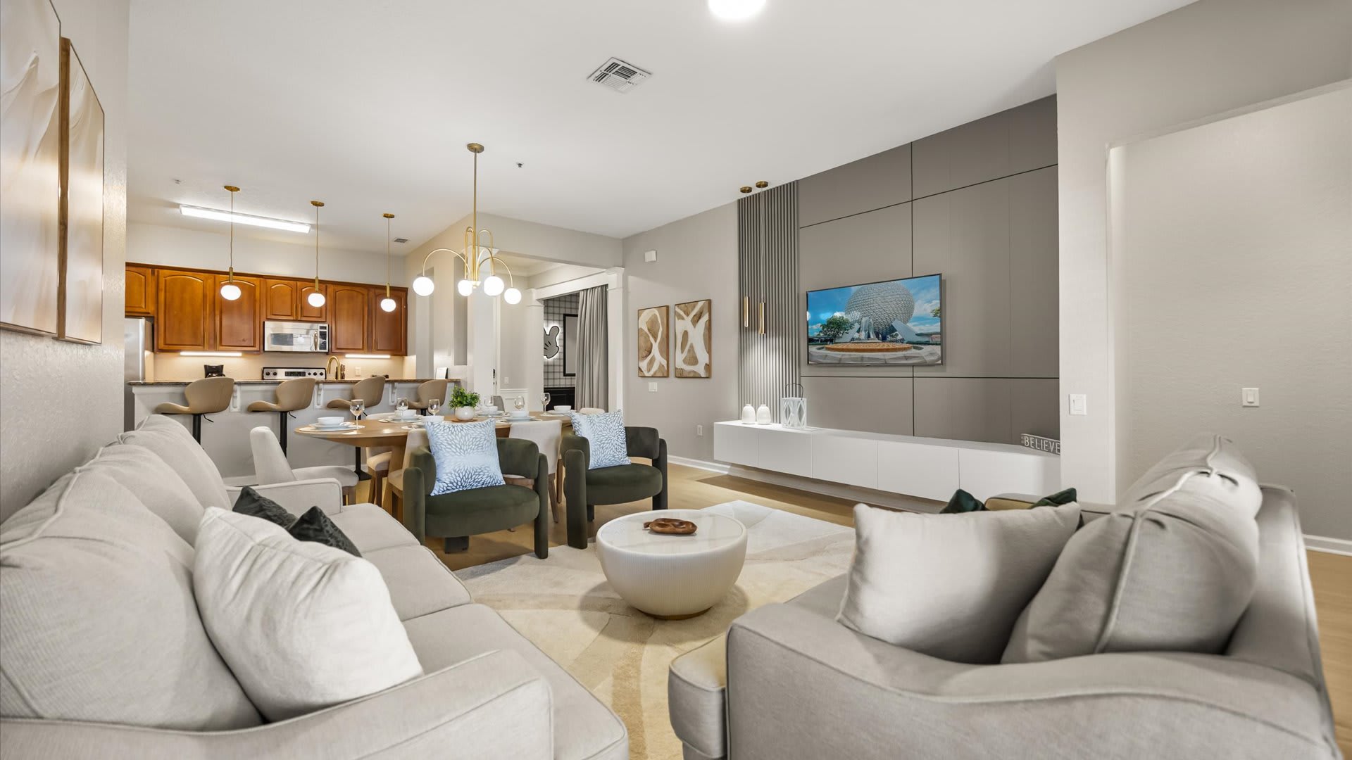 Open-concept living room with a large L-shaped light gray sofa, a round white coffee table, and two dark green armchairs. The dining area with a wood table and a kitchen with a breakfast bar are visible in the background.