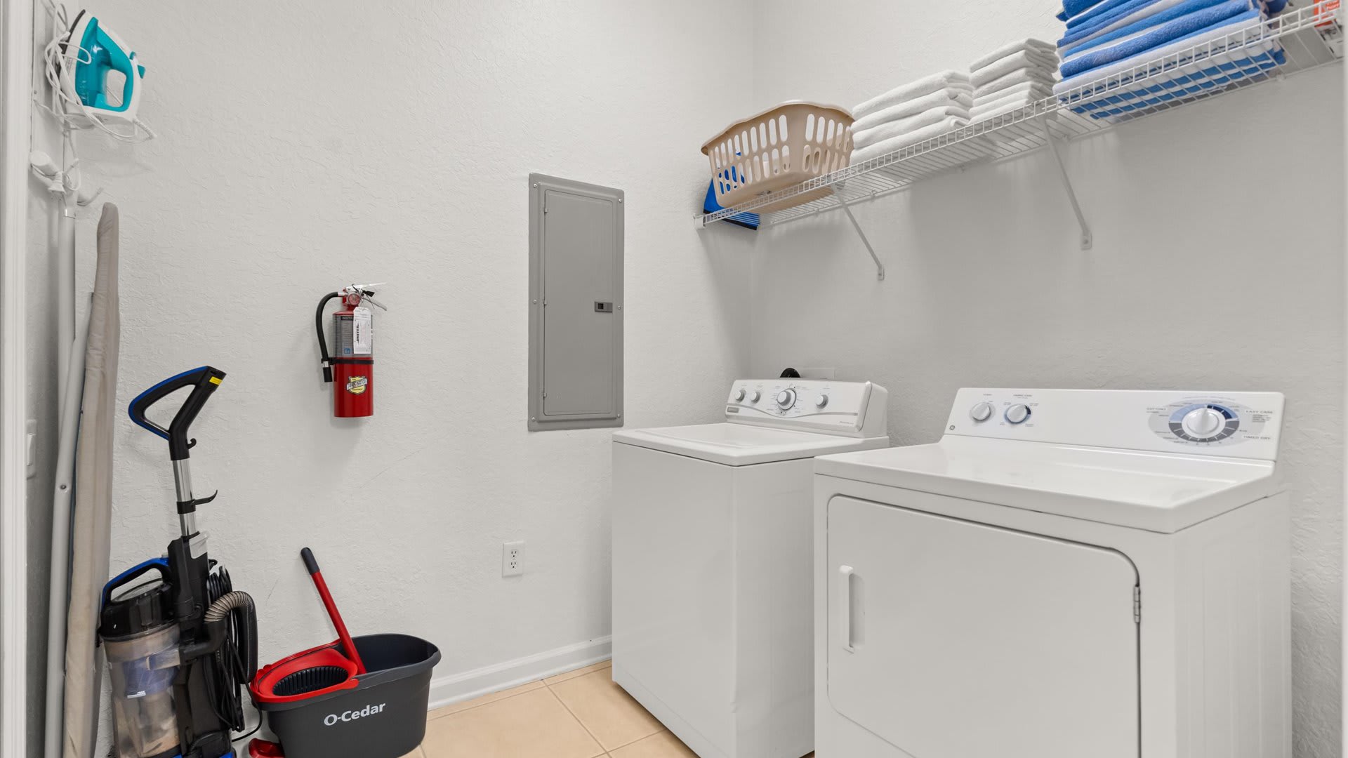 A functional laundry room with a white top-load washing machine and a front-load dryer. A wire shelf above holds folded towels, and cleaning supplies are visible on the light-colored tiled floor.