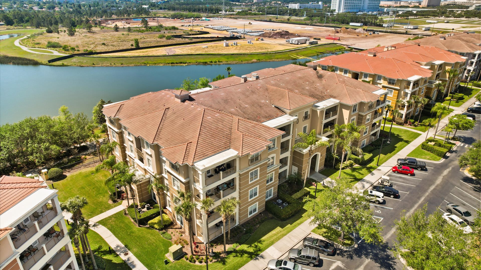 Aerial view of a large, multi-story condominium building with a terracotta tile roof, light-colored facade, and multiple balconies, situated next to a lake with a parking lot in the foreground. Surrounding area shows green landscaping and distant construction.