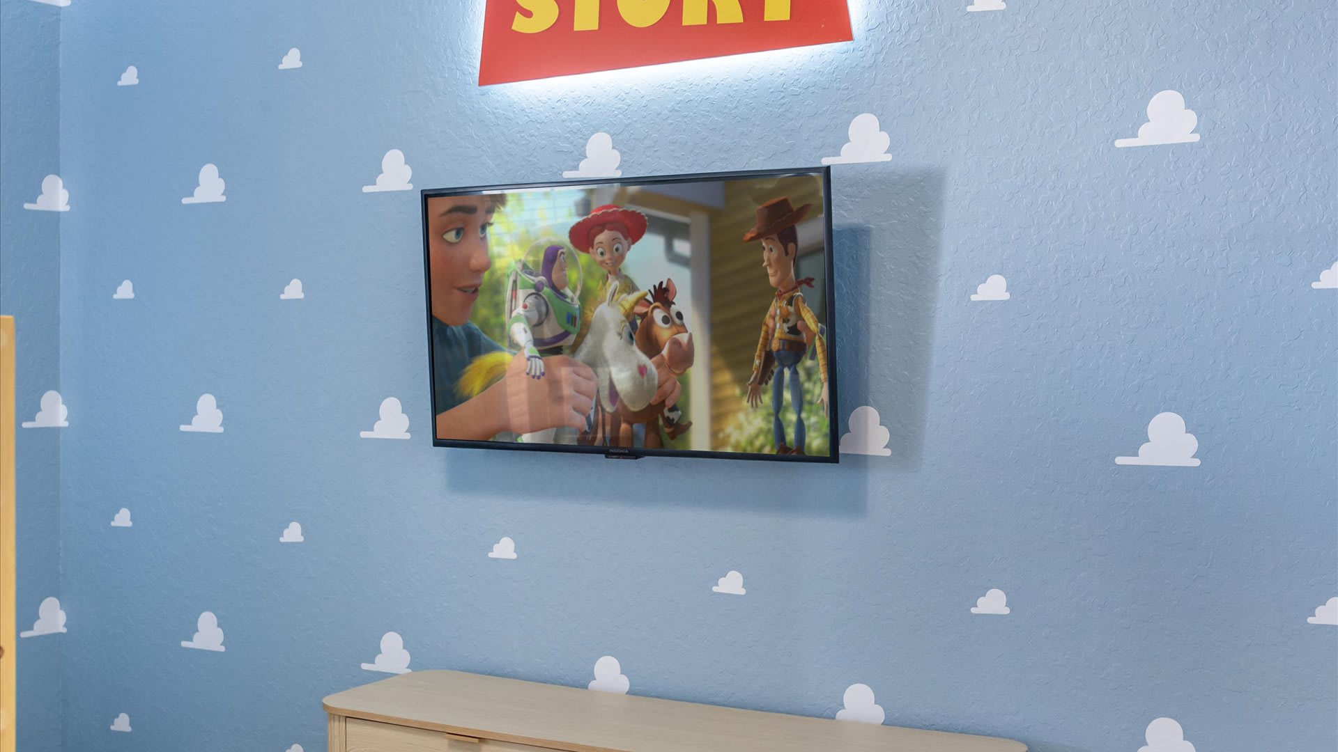 A wall-mounted flat-screen television displaying a scene from Toy Story, set against a light blue wall with white cloud decals. Above the TV, a large illuminated 'TOY STORY' sign is visible.