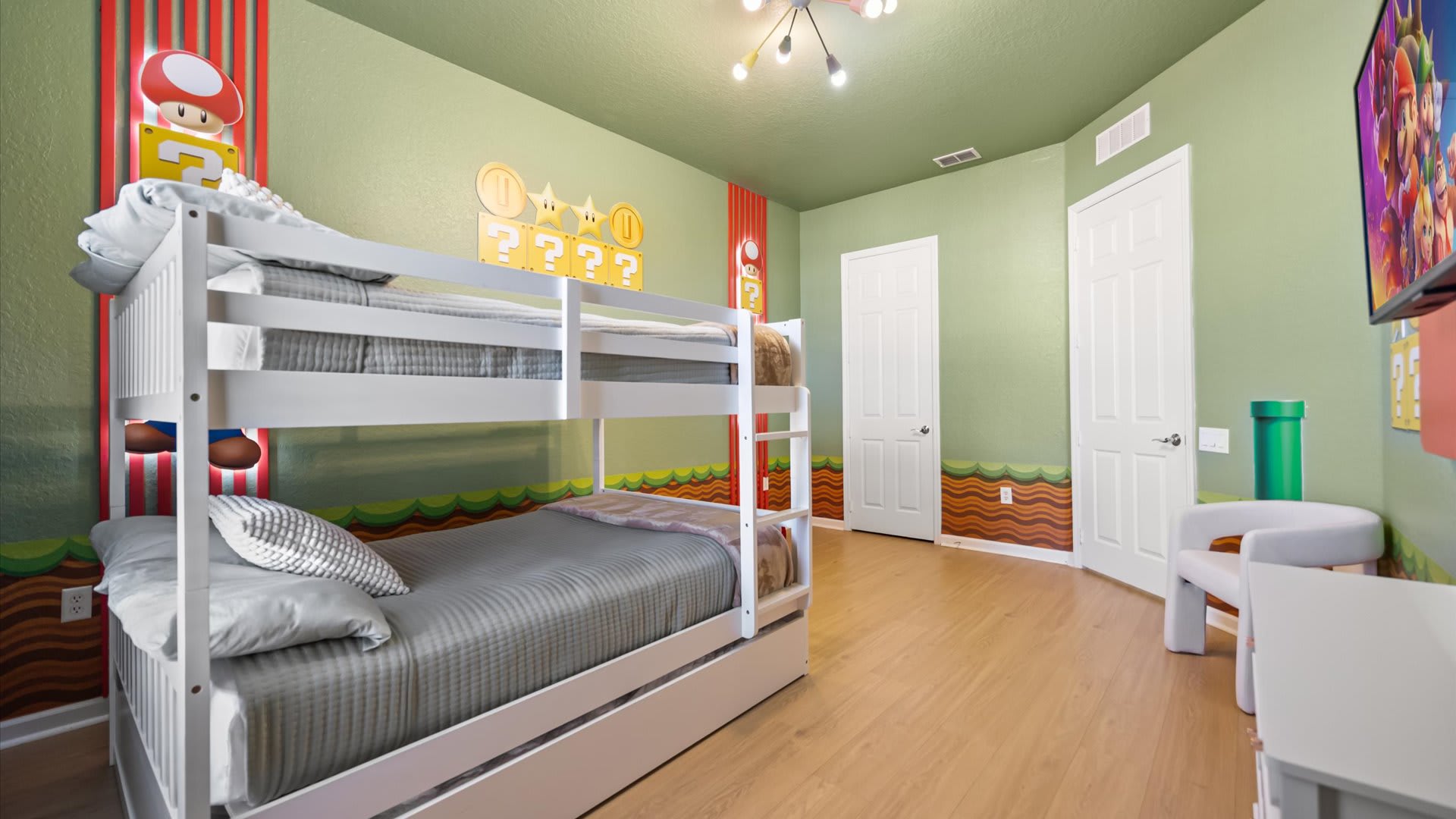 A view of the Super Mario themed bedroom from the opposite corner, showing the white bunk bed with grey bedding and two white doors. The light green walls feature Mario-themed decor and light wood flooring.