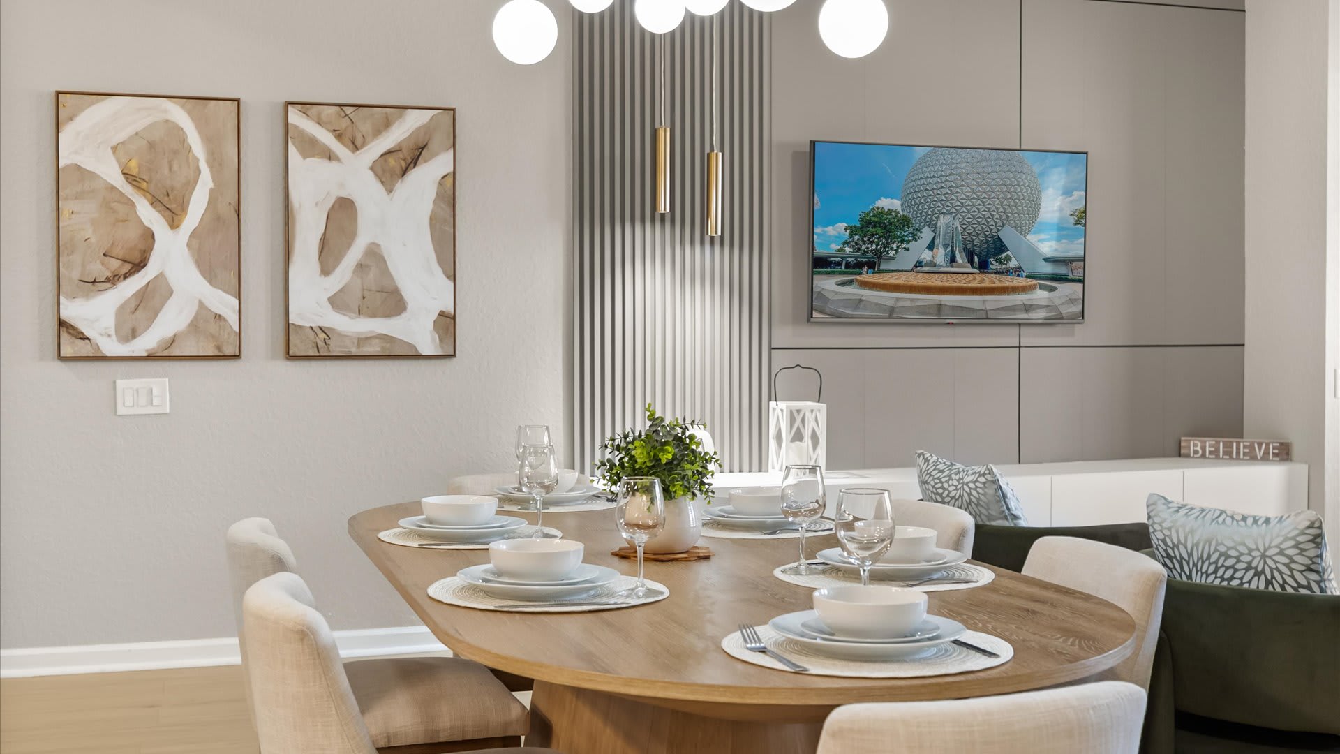 Close-up of the modern dining area with a light wood oval table set for six, featuring cream upholstered chairs and a contemporary gold chandelier. A wall-mounted TV displays a scenic image on a gray accent wall.
