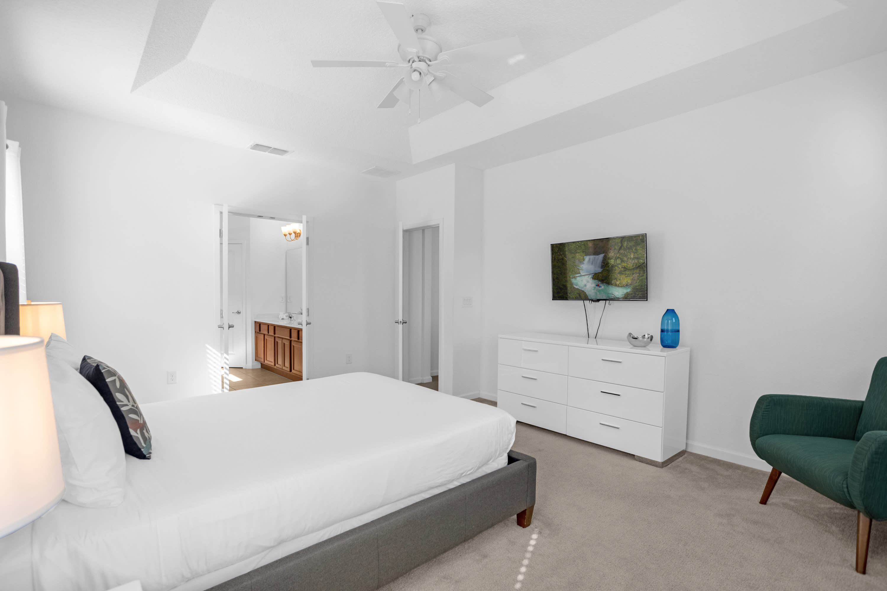 Bright bedroom with white walls and carpet, featuring a king-size bed, a white dresser with a flat-screen TV, and a teal accent chair. The en-suite bathroom entrance is visible.