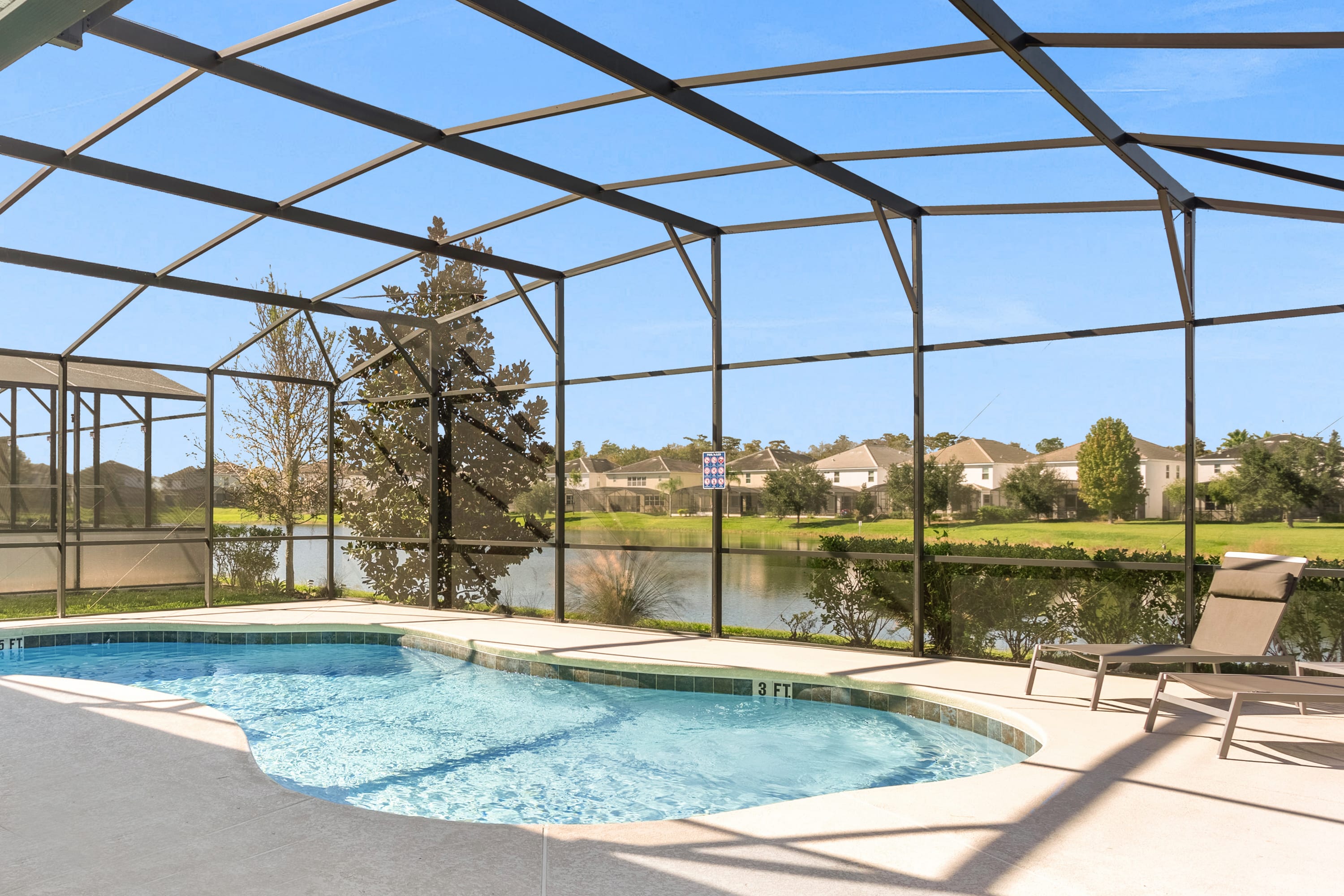An outdoor private pool with light blue water is enclosed by a screened cage, offering a view of a serene lake and residential houses in the distance under a clear blue sky. A lounge chair sits on the light-colored concrete deck.