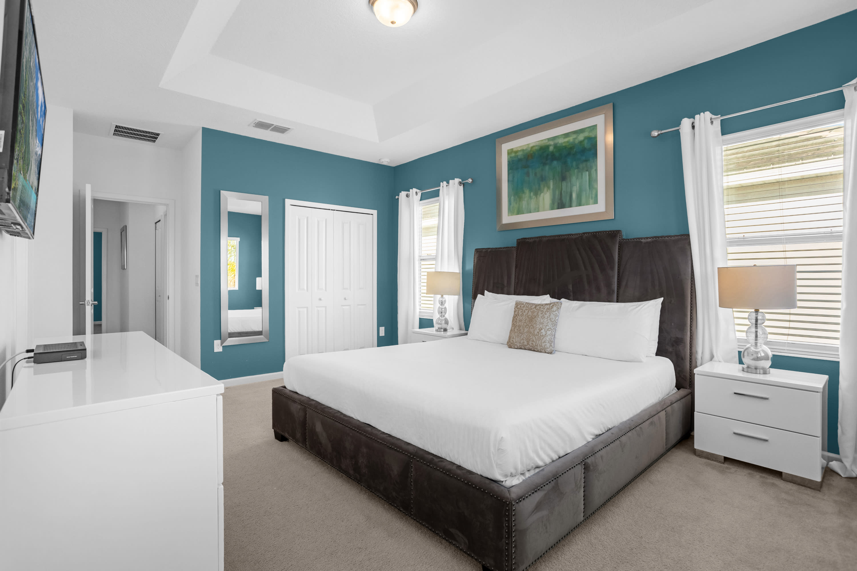 Wide view of a bedroom featuring a king-sized bed with a dark upholstered headboard, white dresser with TV, and a teal accent wall. A full-length mirror is visible next to a closet.