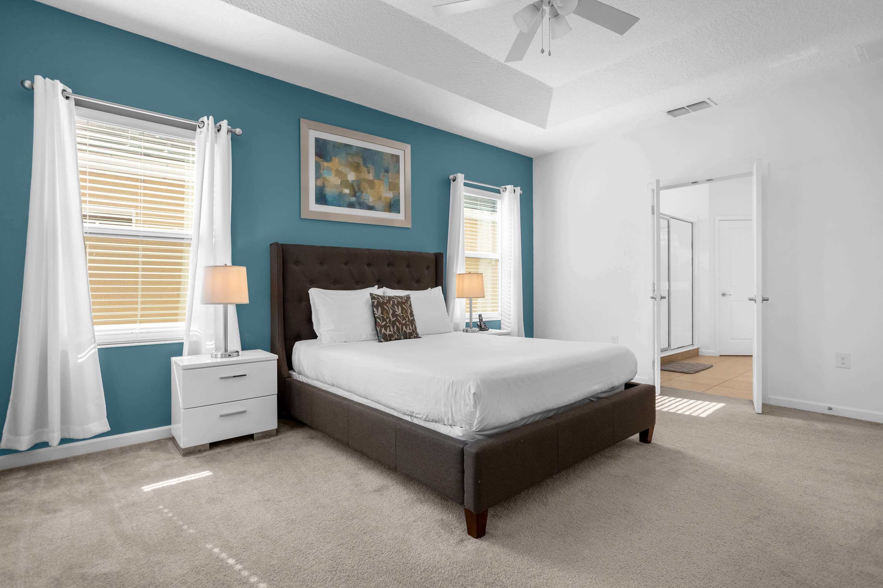 Bright bedroom with a king-sized bed, white nightstands, and a teal accent wall with two windows. An open doorway leads to an en-suite bathroom.