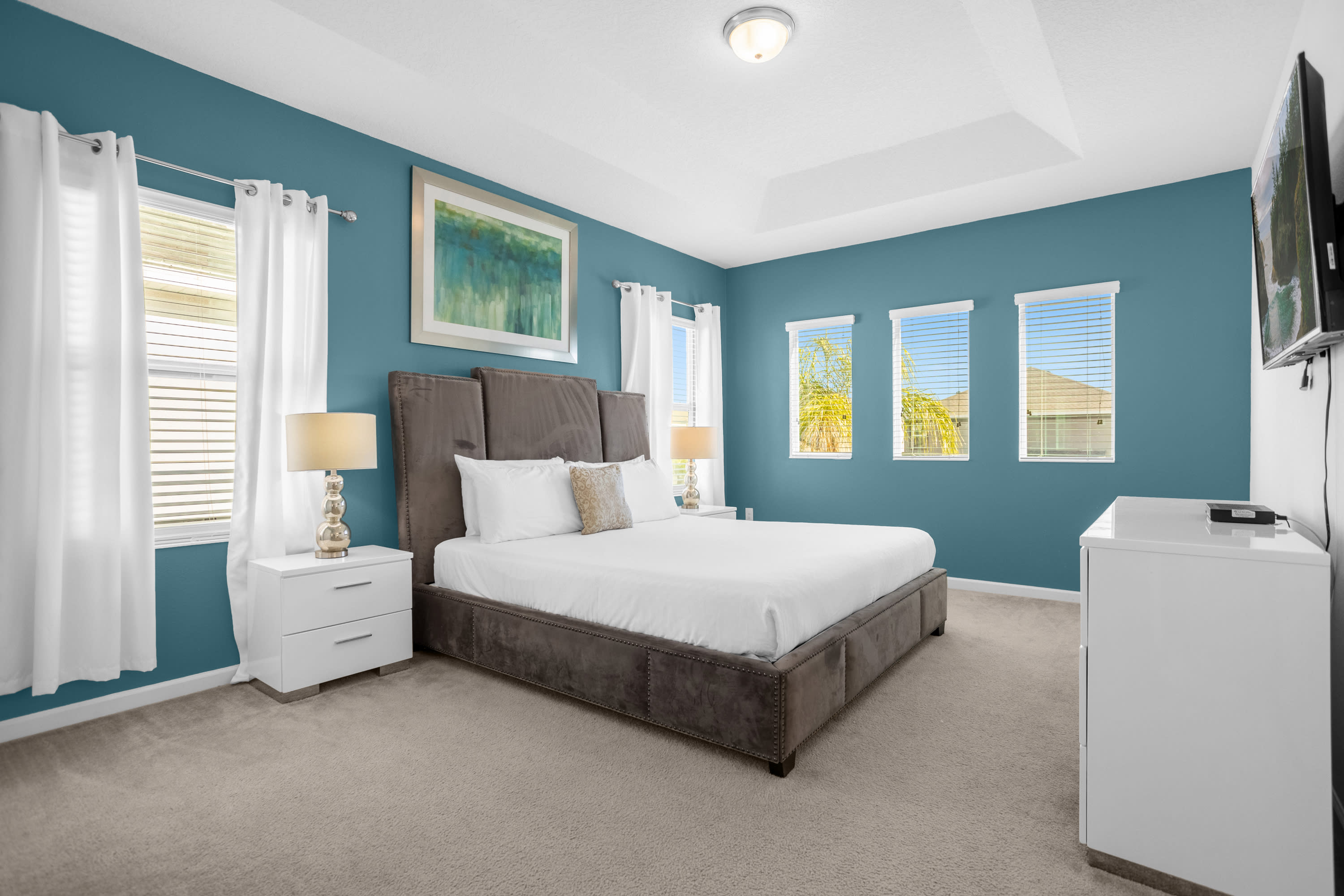 Spacious master bedroom with a teal accent wall and white walls, featuring a large king-sized bed with a tall upholstered headboard. White nightstands with lamps flank the bed, and multiple windows with white curtains provide ample light.