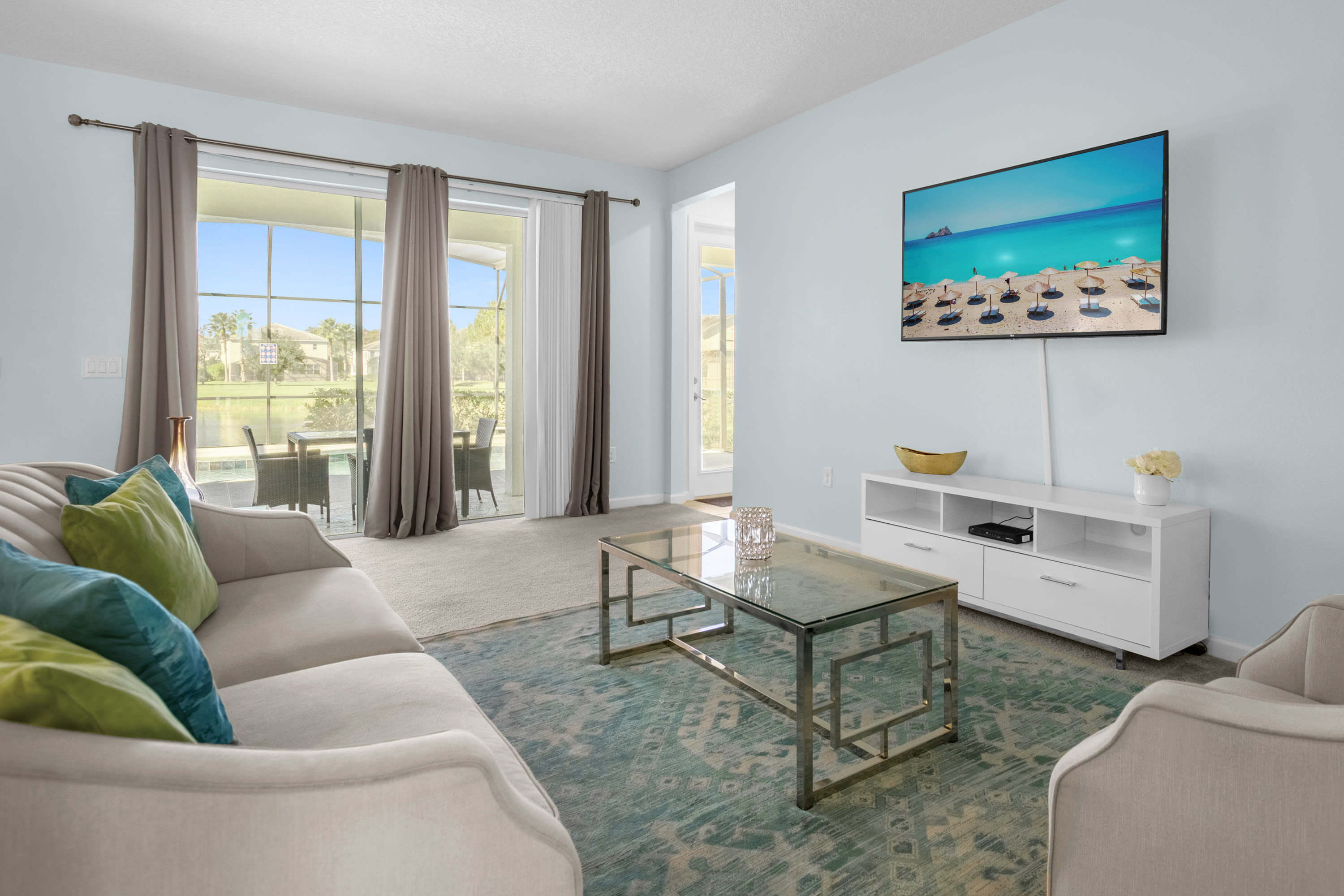 A bright living room with light blue walls features a wall-mounted flat-screen TV above a white media console. A beige sofa and a glass coffee table sit on a blue patterned rug, with sliding glass doors offering a view of the outdoor area.