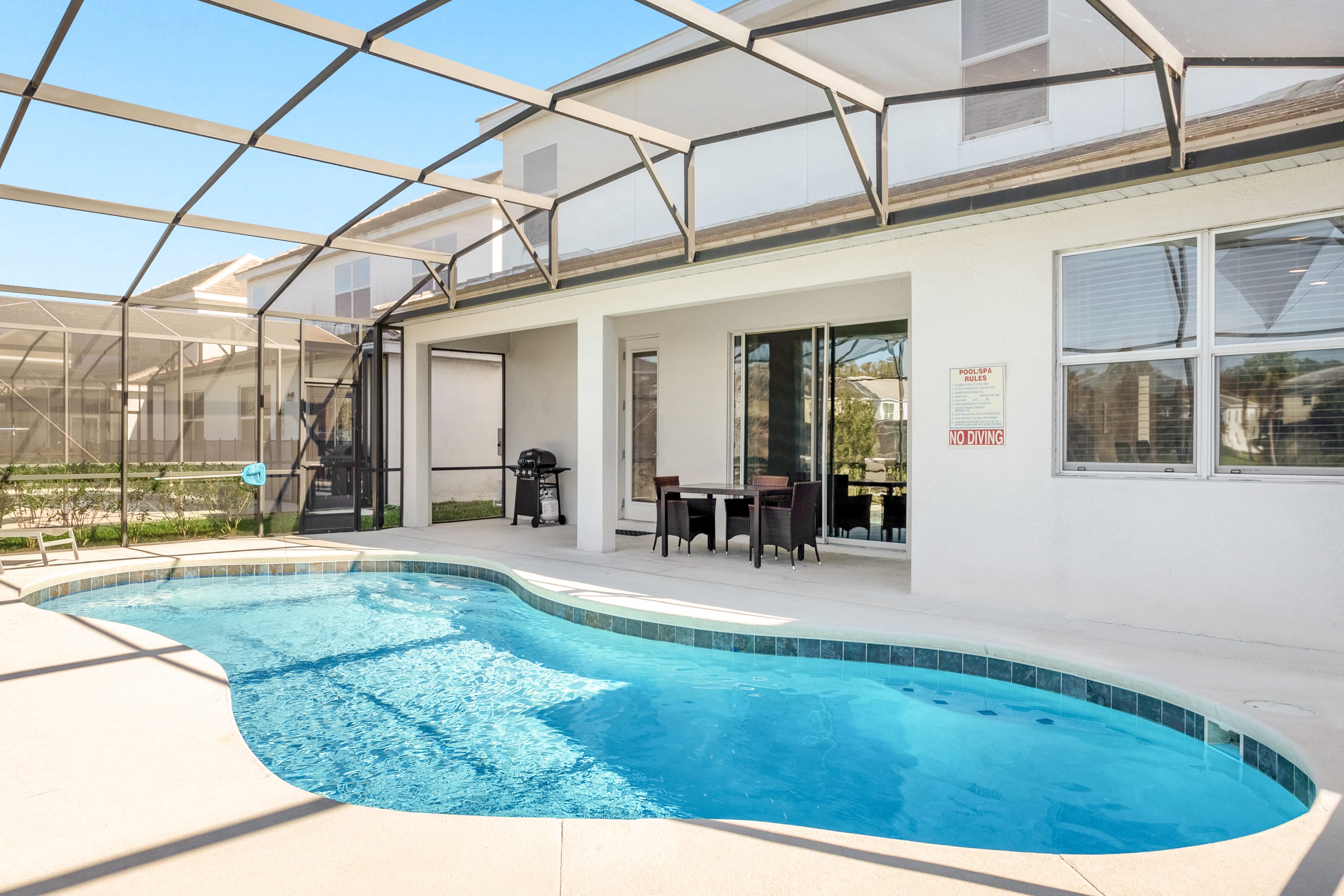 A covered patio area next to the private pool features an outdoor dining table with chairs and a BBQ grill. The white exterior of the house with large windows and sliding glass doors opens to the screened pool deck.