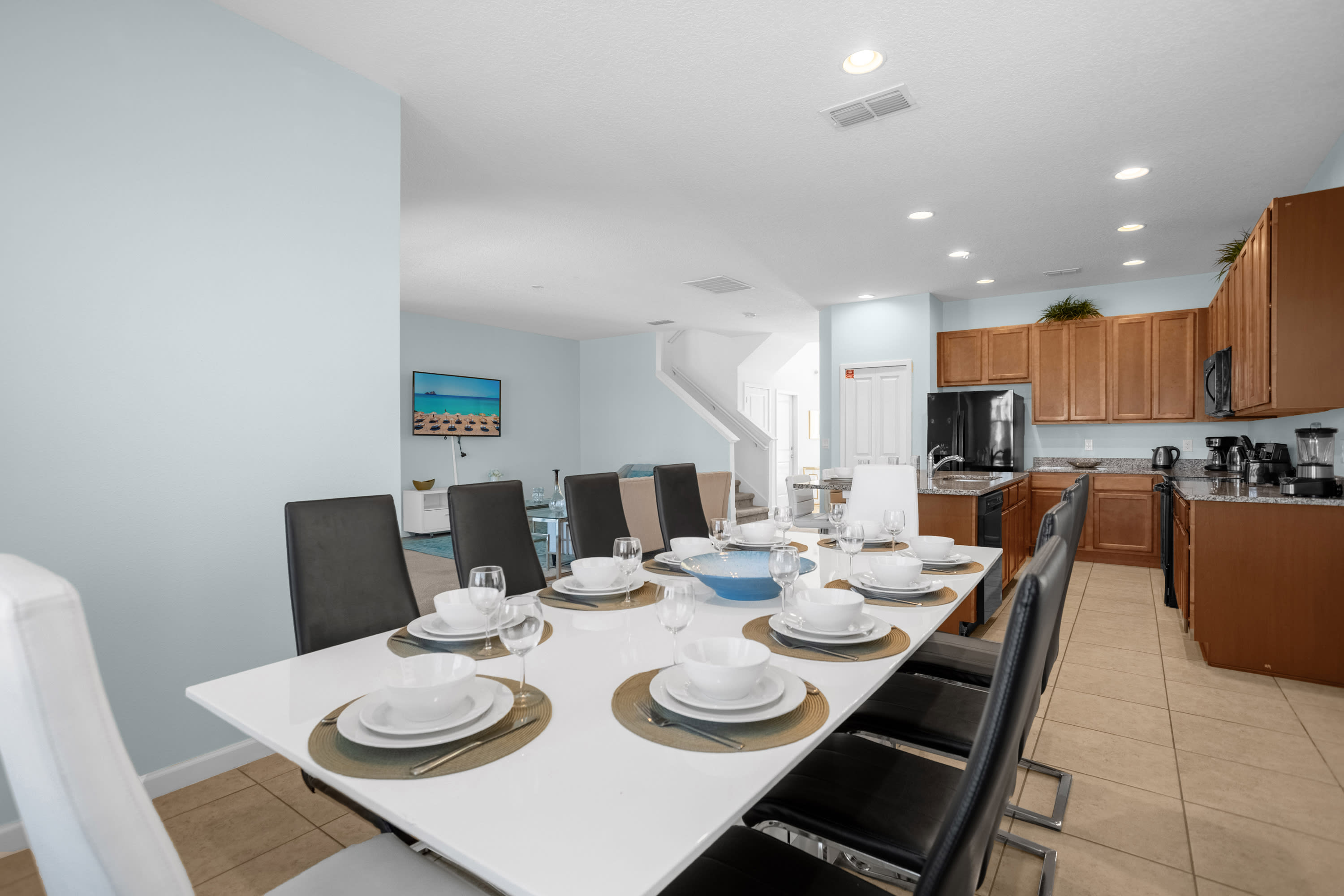 Spacious dining area featuring a long white table set with dinnerware and black and white chairs, with the kitchen and living room visible in the background. Light blue walls and tile flooring complete the space.