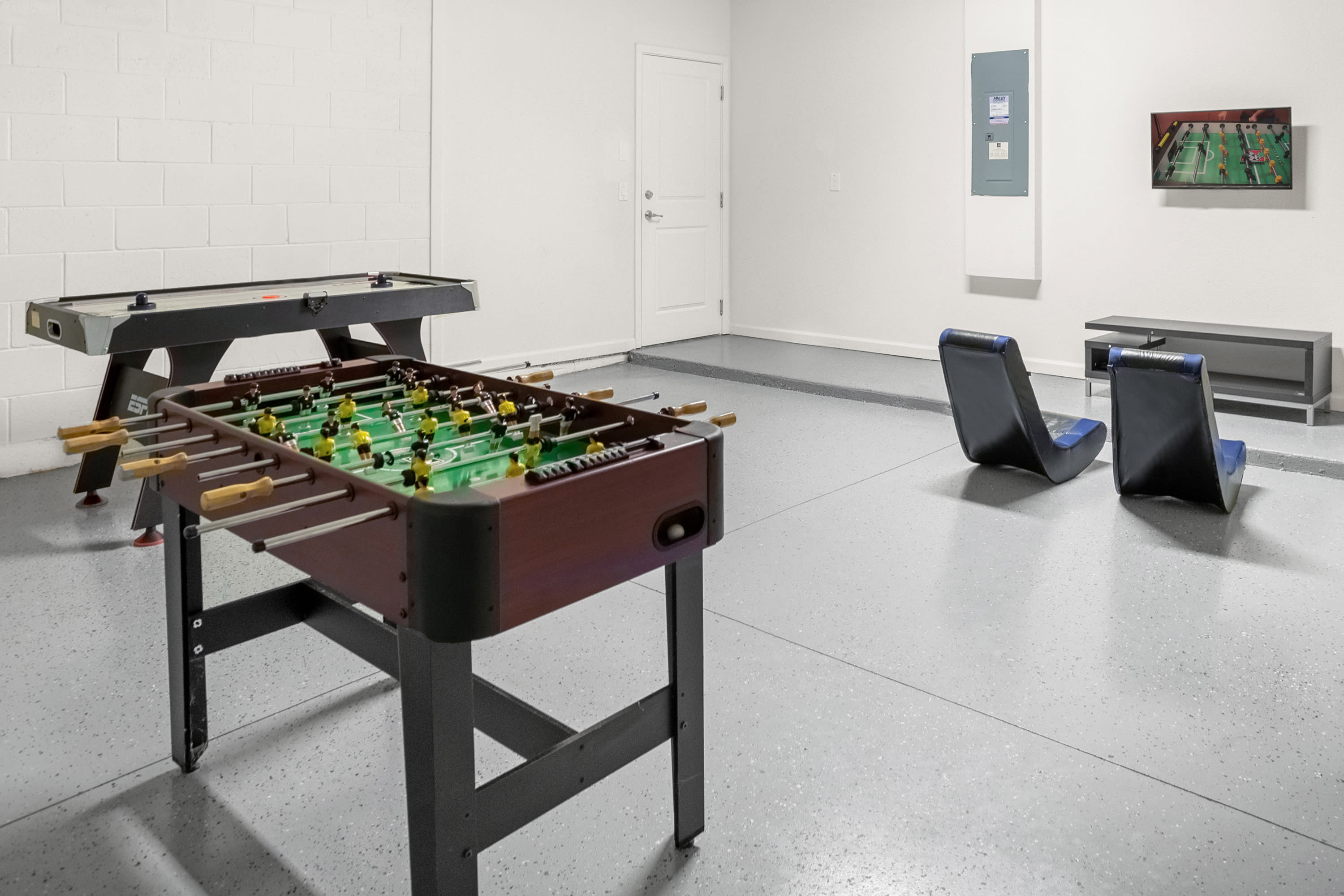 Another view of the game room shows a foosball table and an air hockey table on a grey speckled floor. White walls and a white door are visible, with two low-profile chairs and a small TV in the background.