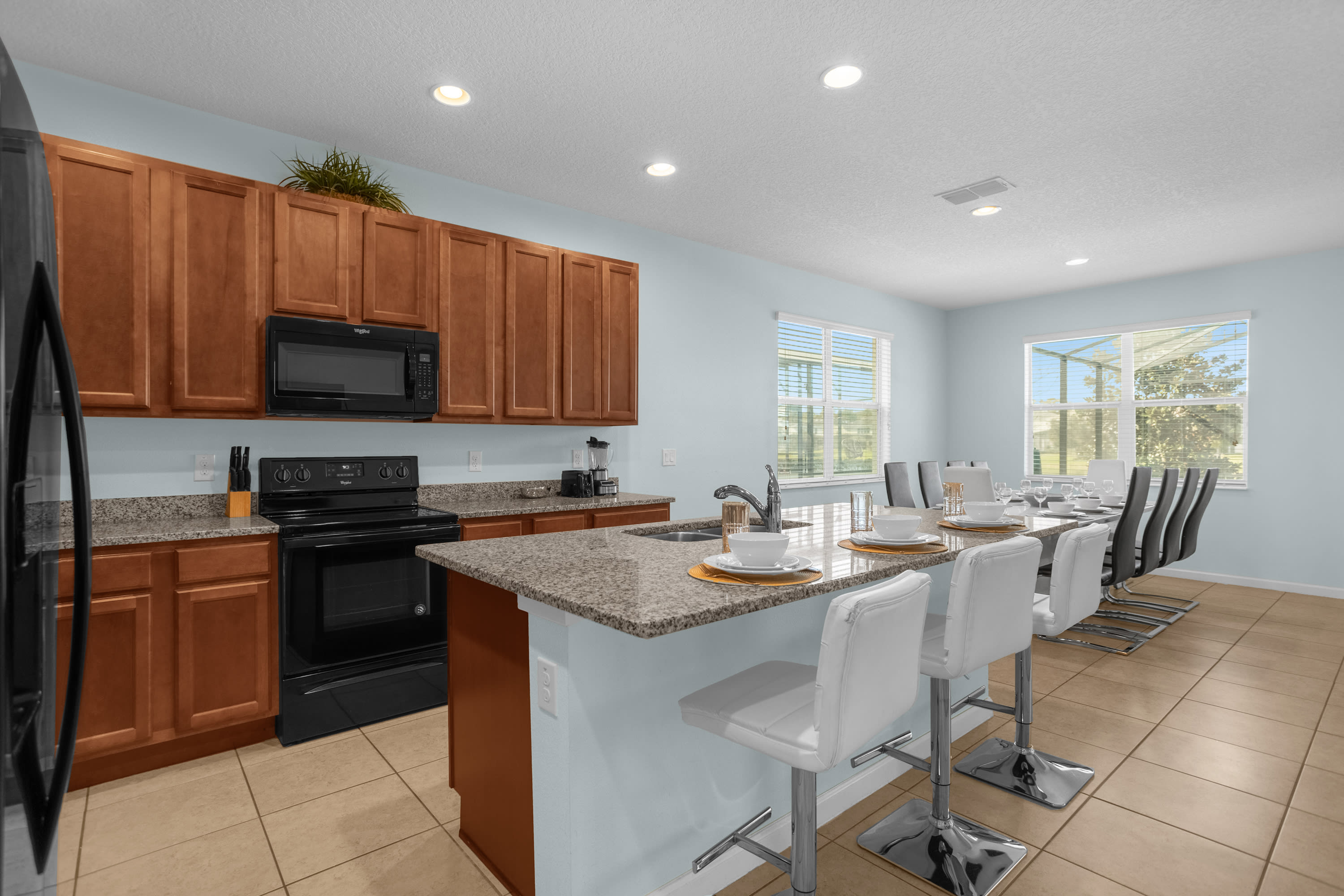 Contemporary kitchen with light blue walls, wood cabinets, and granite countertops, featuring a black electric stove and a central island with three white bar stools. The dining area is visible through the windows.
