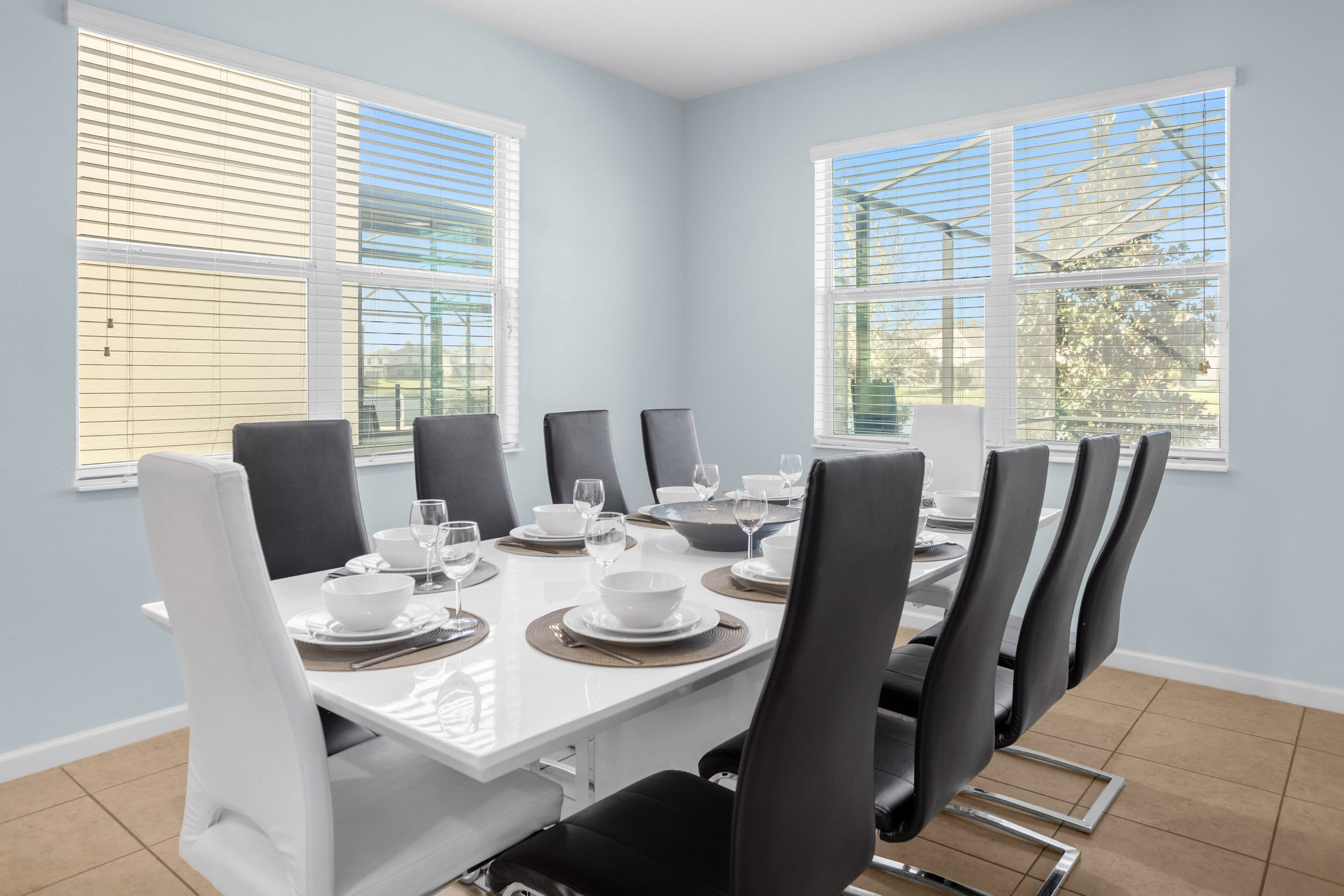 Elegant dining room with a large white table set for multiple guests, surrounded by black and white chairs. Two windows with blinds offer views of the outdoor patio area.
