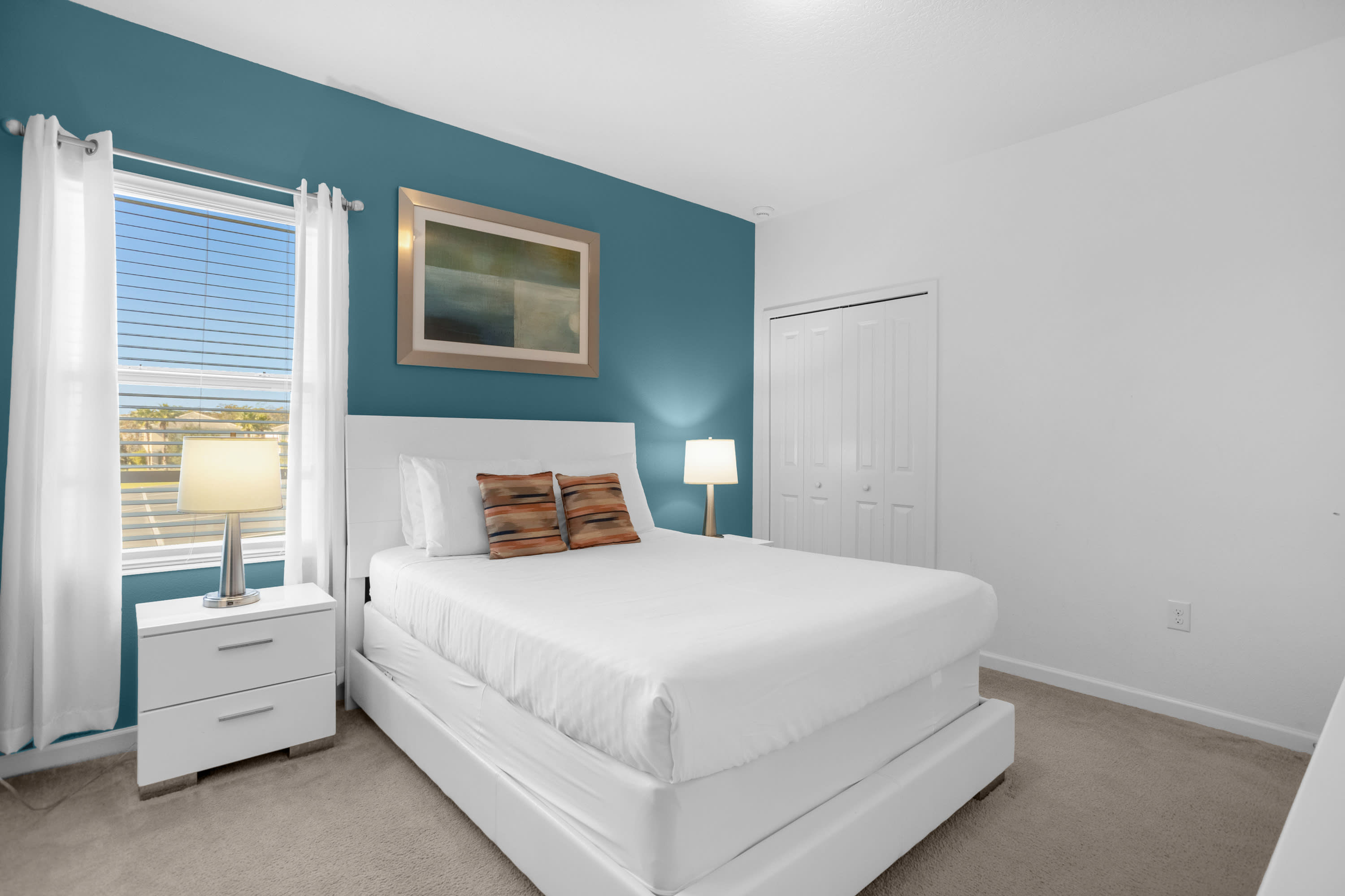 Bright bedroom featuring a white queen-sized bed with white bedding and orange accent pillows, flanked by white nightstands with lamps. A teal accent wall and a window with blinds are visible.