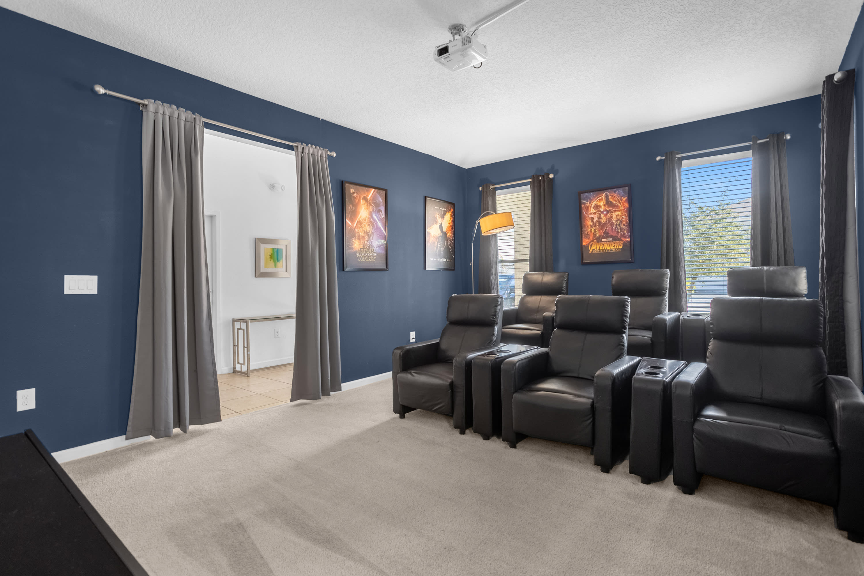 A wide view of the media room shows multiple black leather recliners arranged in rows, facing a large projection screen. Dark blue walls are adorned with movie posters, and heavy grey curtains frame the windows.
