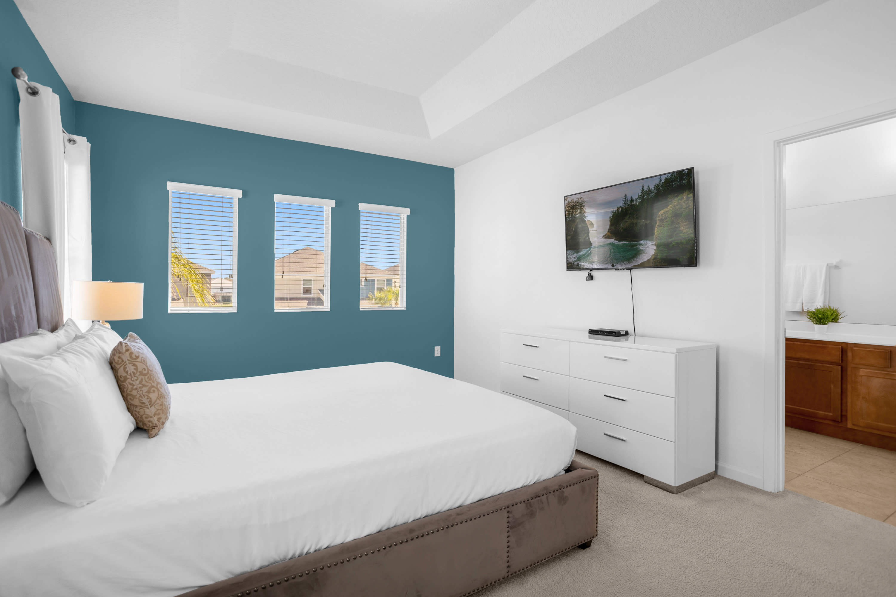 Bedroom with a king-sized bed, a white dresser with a wall-mounted TV, and a teal accent wall featuring three windows with blinds. An open doorway leads to an en-suite bathroom.