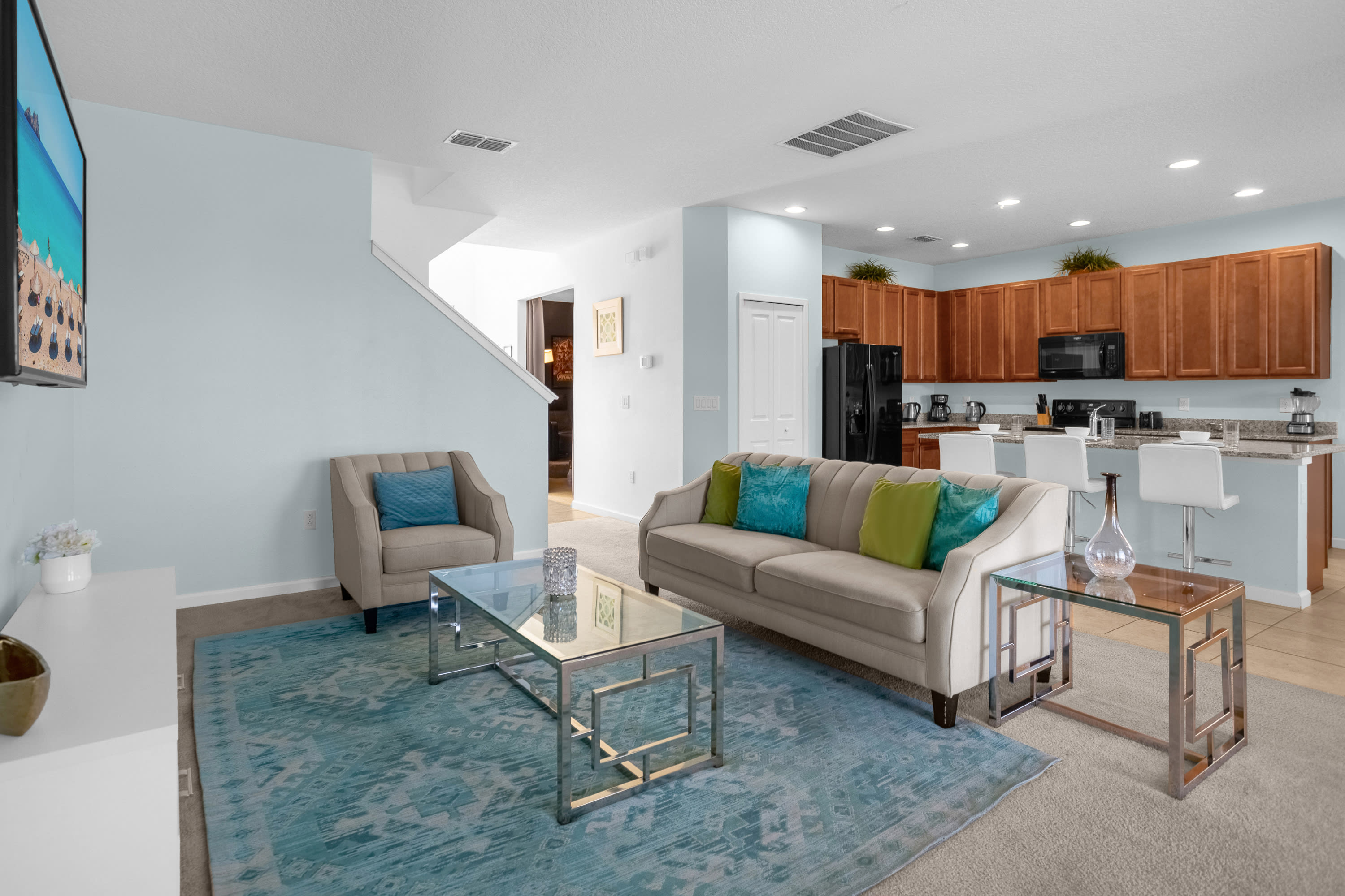 A bright living room features light blue walls, a beige sofa with blue and green throw pillows, and a glass coffee table on a patterned blue rug. The open-concept space connects to a kitchen with wood cabinets and a breakfast bar.
