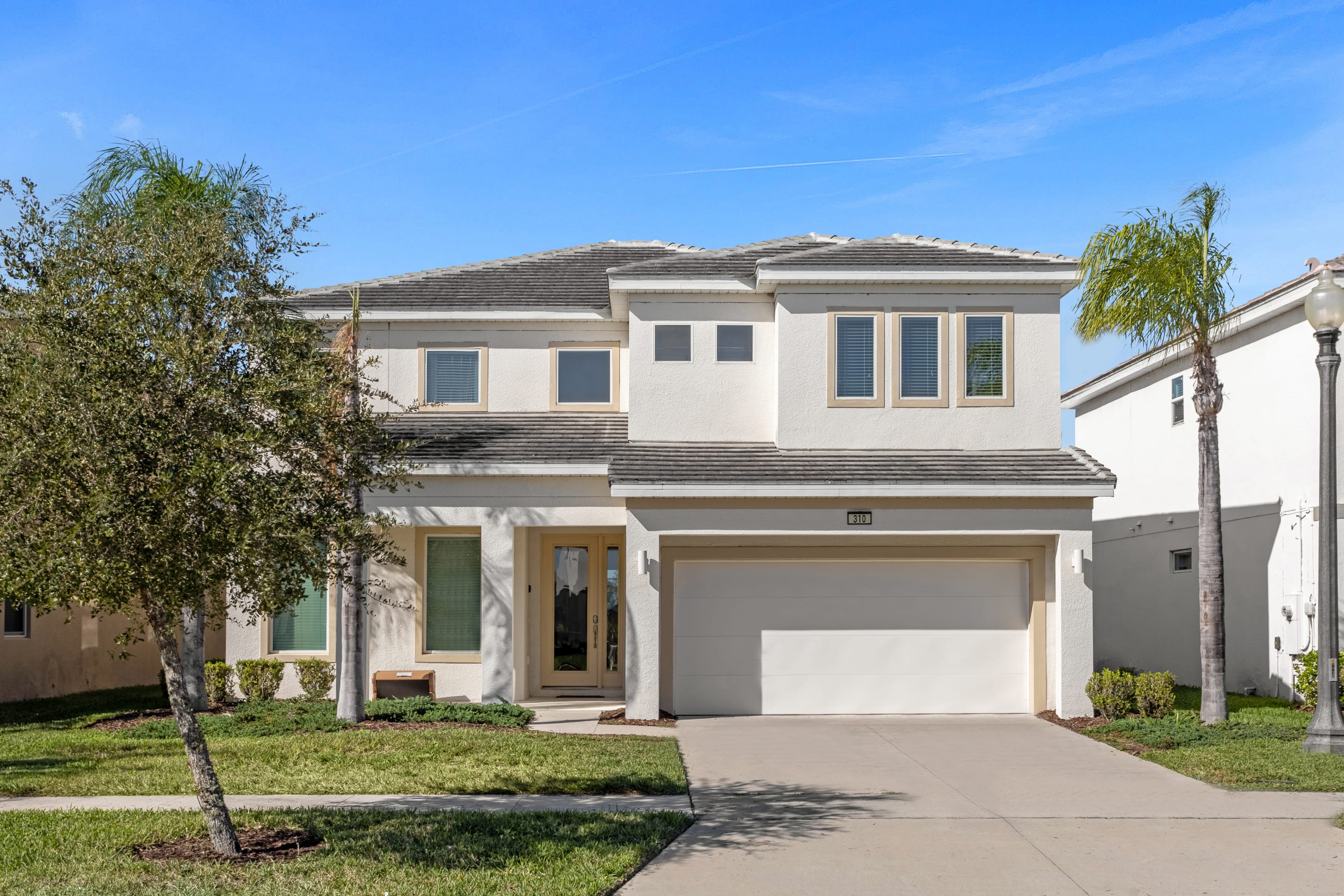 A modern two-story house with a light stucco exterior and a dark tile roof features multiple windows, a two-car garage, and a paved driveway, flanked by green lawns and palm trees under a clear blue sky.