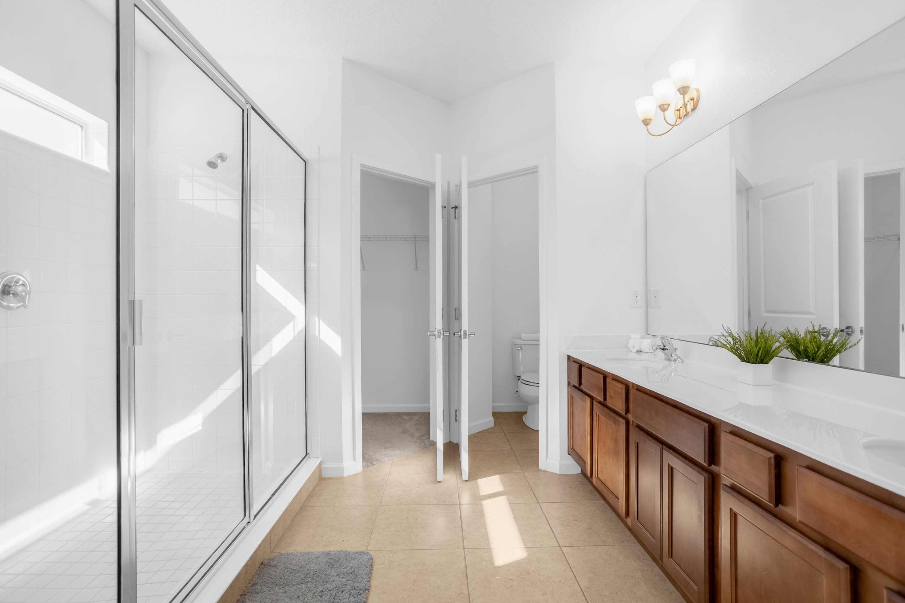 Spacious bathroom with a walk-in shower, double vanity, and a separate toilet area with bi-fold closet doors. The room features white walls and light tile flooring.