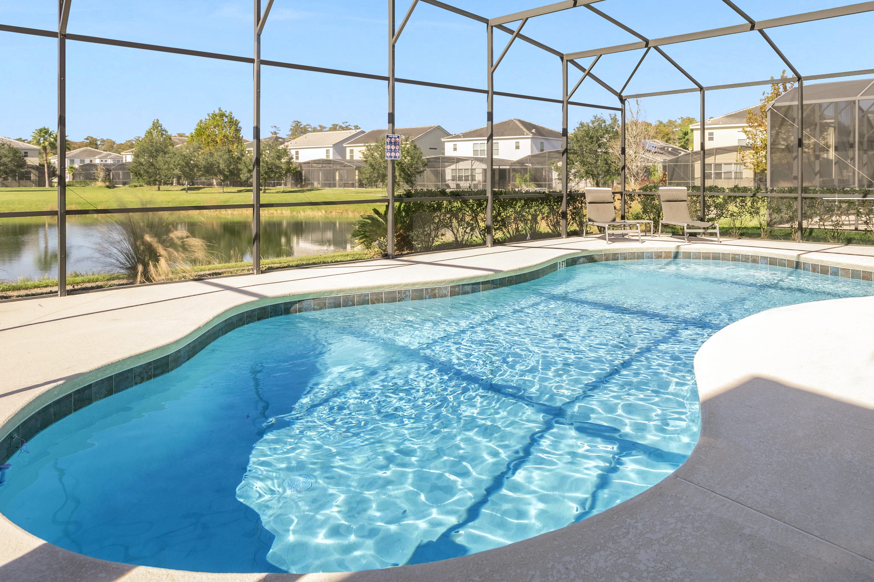 A private outdoor pool with clear blue water is surrounded by a light concrete deck and enclosed by a screened cage. Beyond the screen, a tranquil lake and residential homes are visible under a bright blue sky.