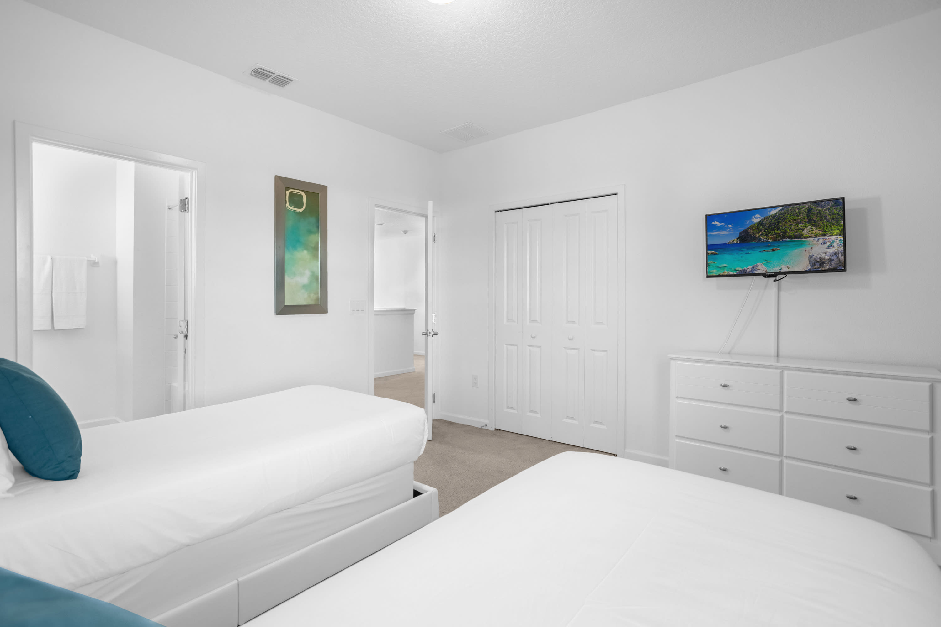 Clean white bedroom featuring two white twin beds, a white dresser with a TV, and a closet with bi-fold doors. A doorway leads to an adjacent bathroom, and abstract artwork hangs on the wall.