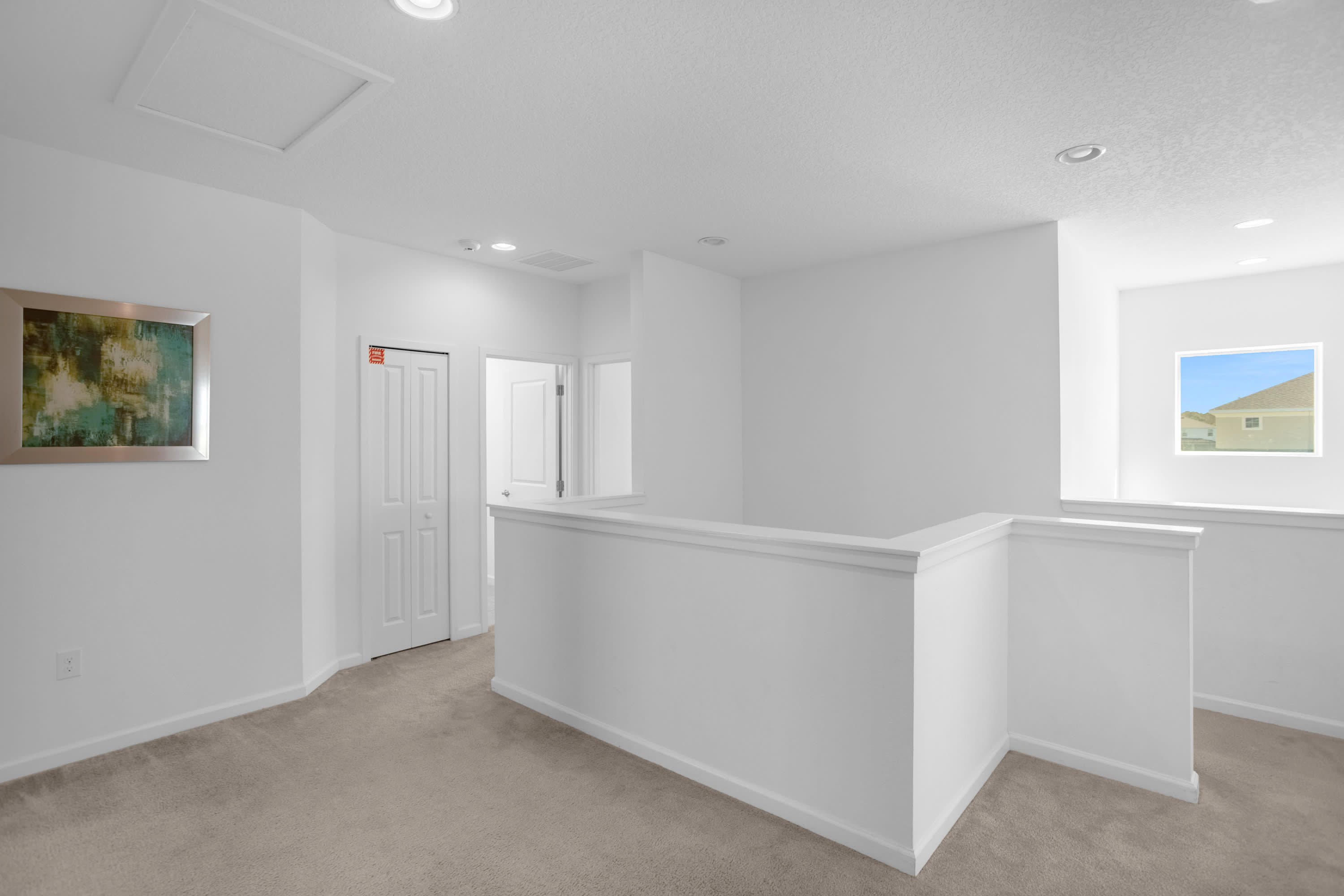 Bright, open loft area with light beige carpet, white walls, and a white railing overlooking an open space. A closed door and a window are visible in the background.