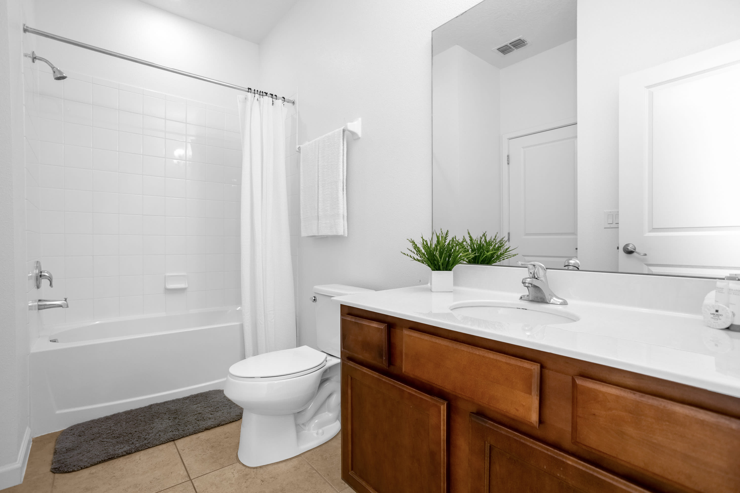 Clean white bathroom with a toilet, a bathtub with a shower and white curtain, and a wooden vanity with a white countertop and sink. A small green plant adds a touch of freshness to the counter.