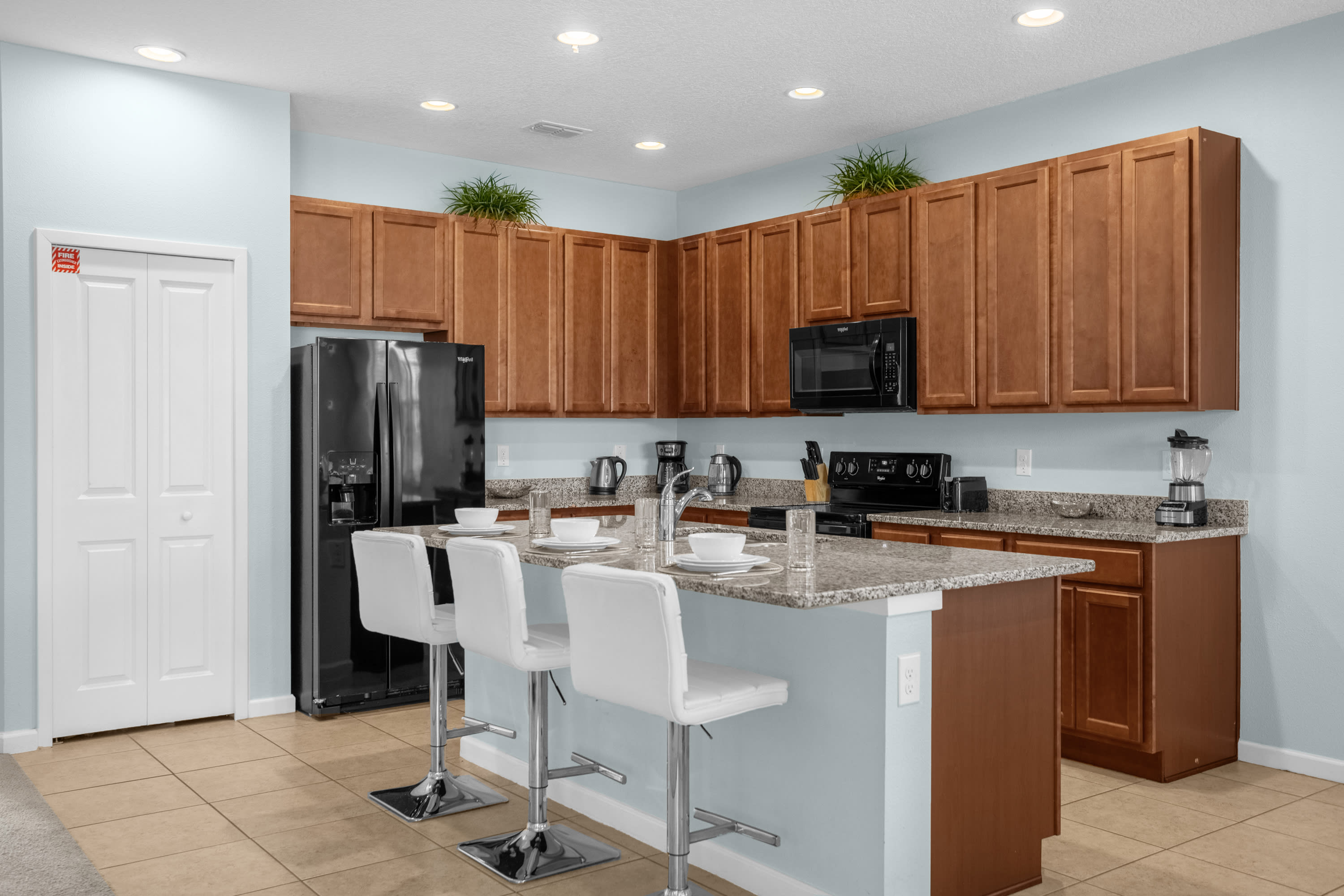 Modern kitchen featuring light blue walls, wood cabinetry, granite countertops, and a central island with three white bar stools. A black refrigerator and microwave are visible.