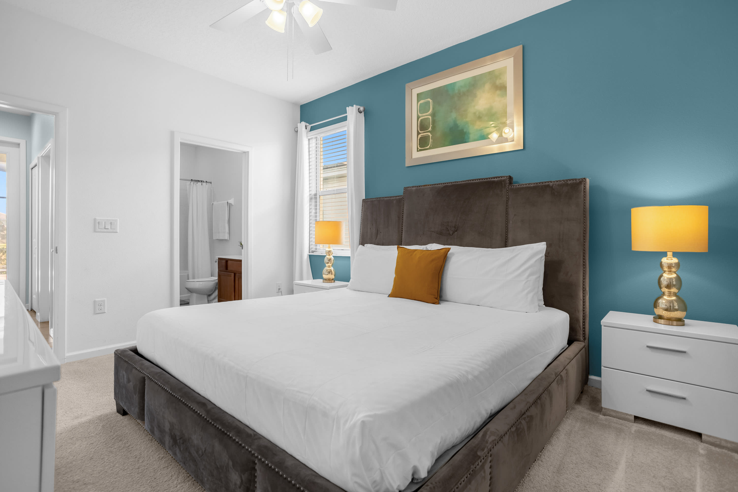 Spacious bedroom with a king-sized bed featuring a tall, dark upholstered headboard, white bedding, and an accent wall painted in a deep teal color. Two white nightstands with yellow lamps flank the bed.