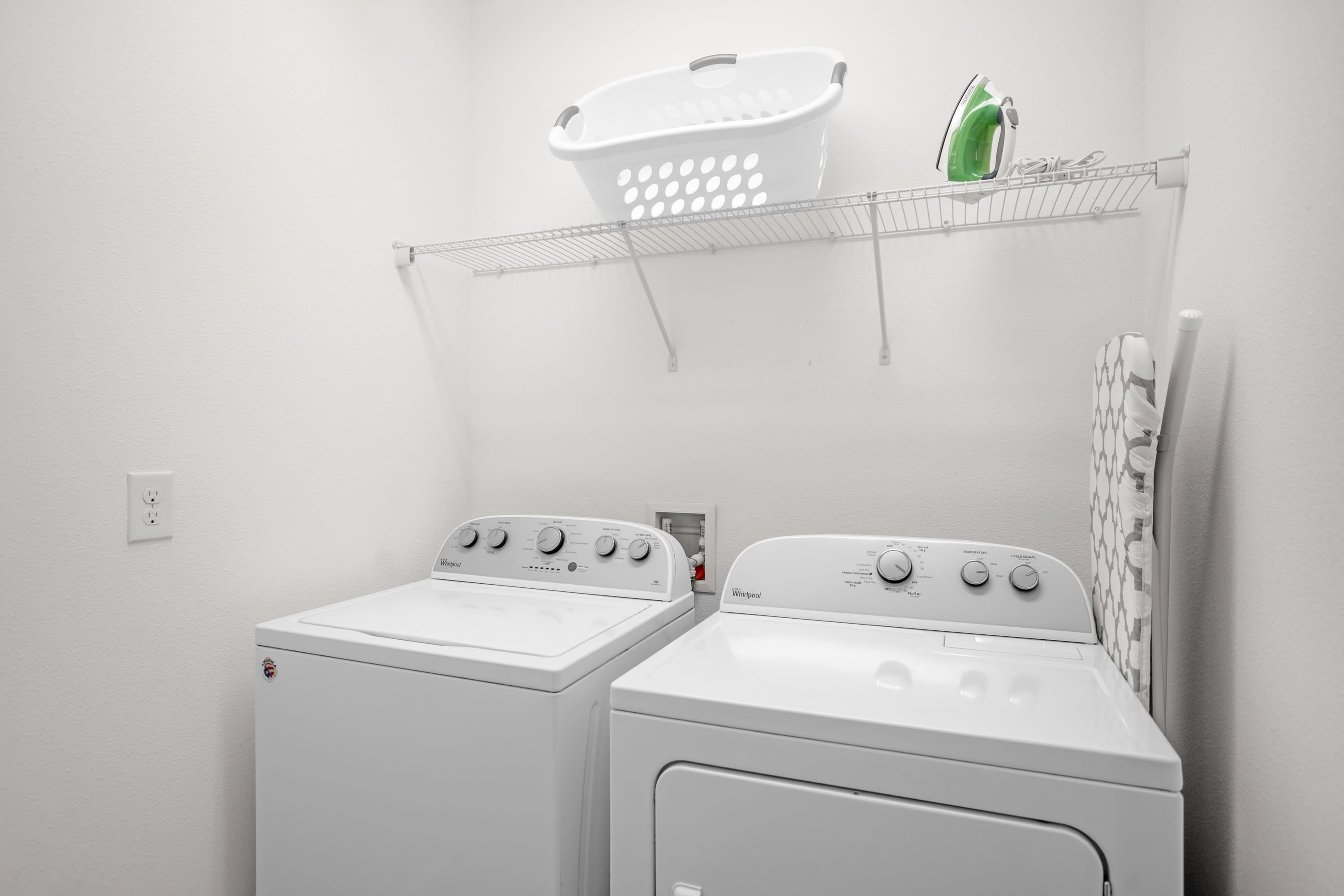 A bright white laundry room features a top-loading washing machine and a front-loading dryer, both white, beneath a wire shelf holding a white laundry basket and a green iron.