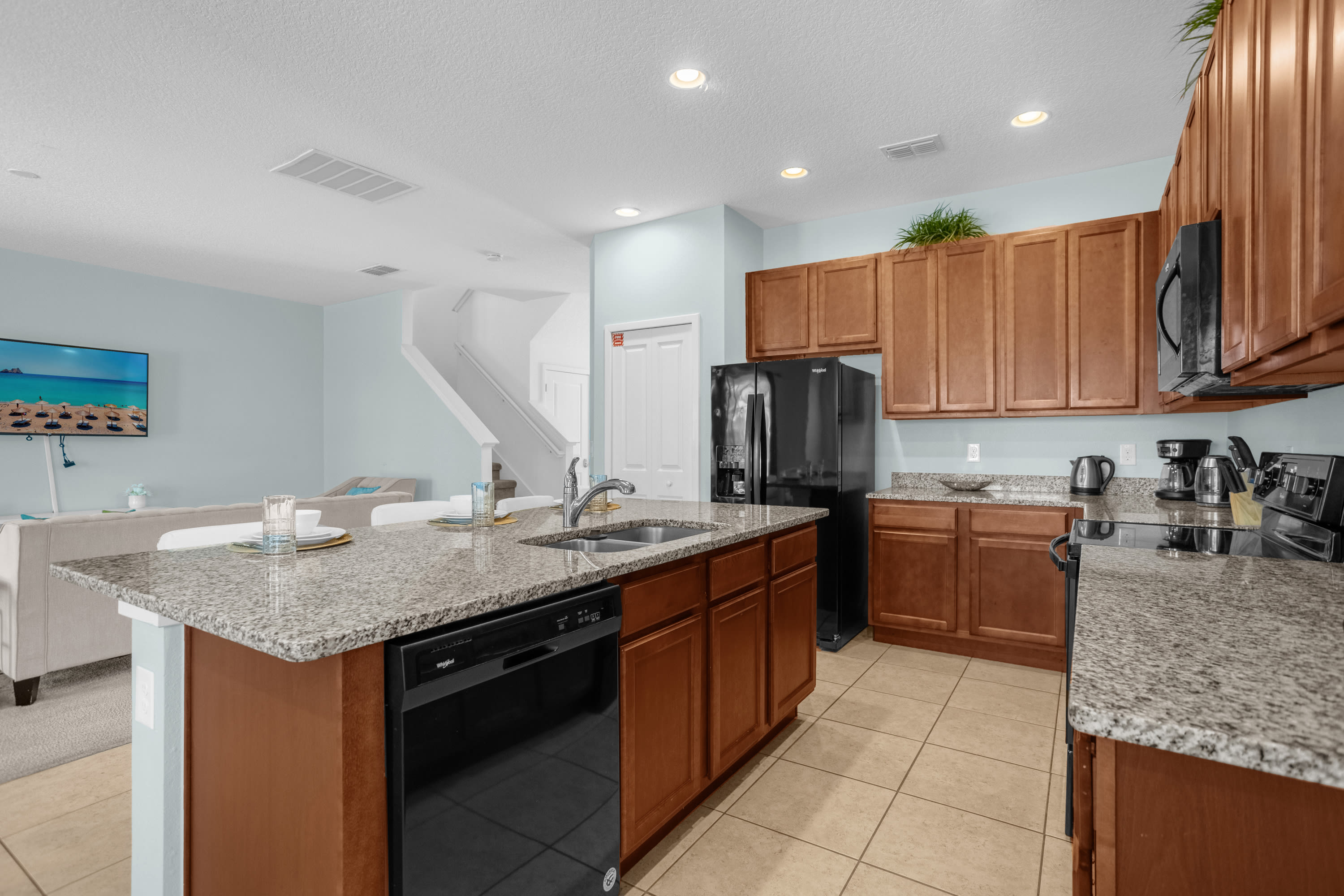 Bright kitchen with light blue walls, wood cabinets, and granite countertops, centered around an island with a sink and black dishwasher. The living room is visible in the background.