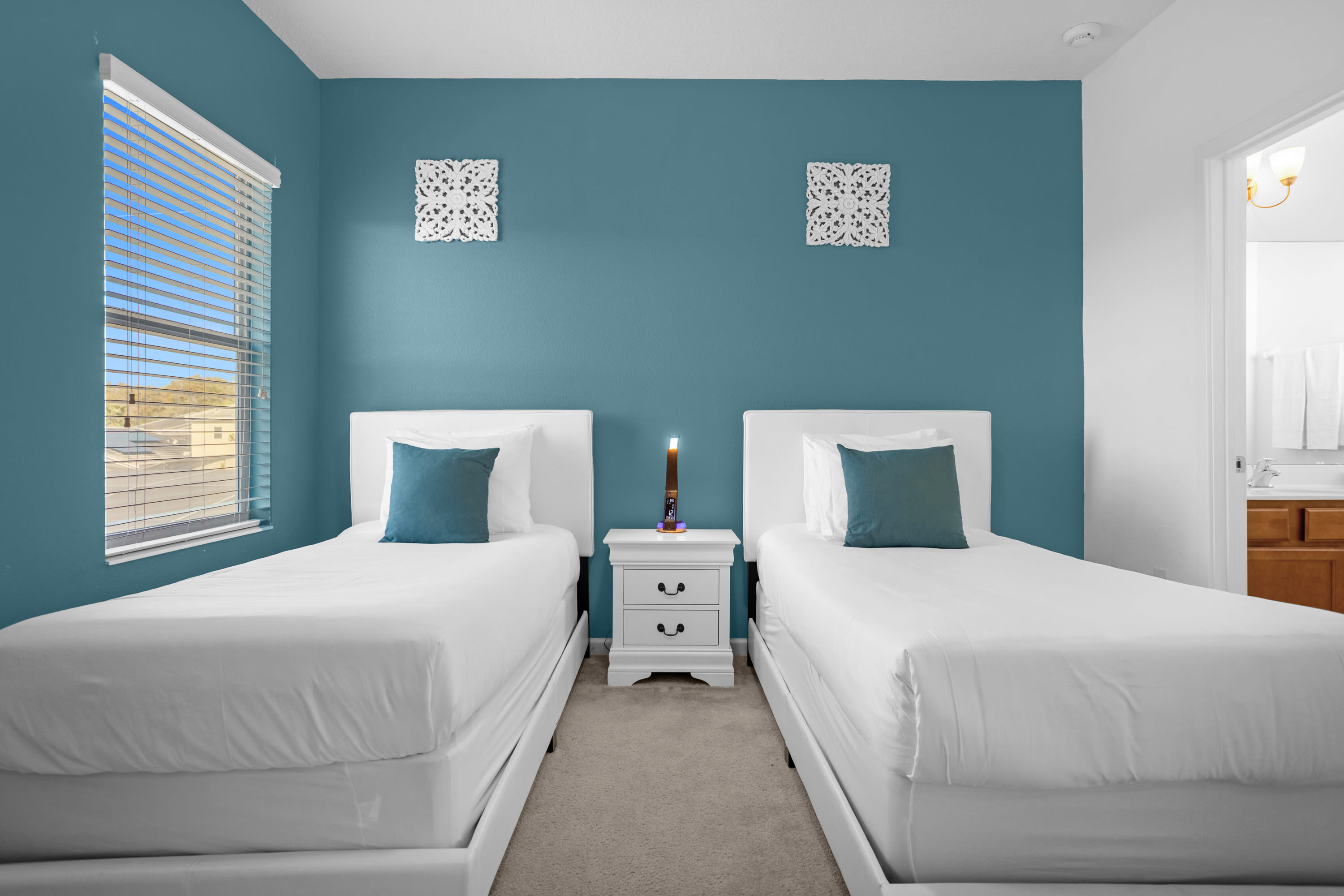 Kids' bedroom featuring two white twin beds with teal accent pillows, separated by a white nightstand with a decorative candle. A teal accent wall and a window with blinds are visible.